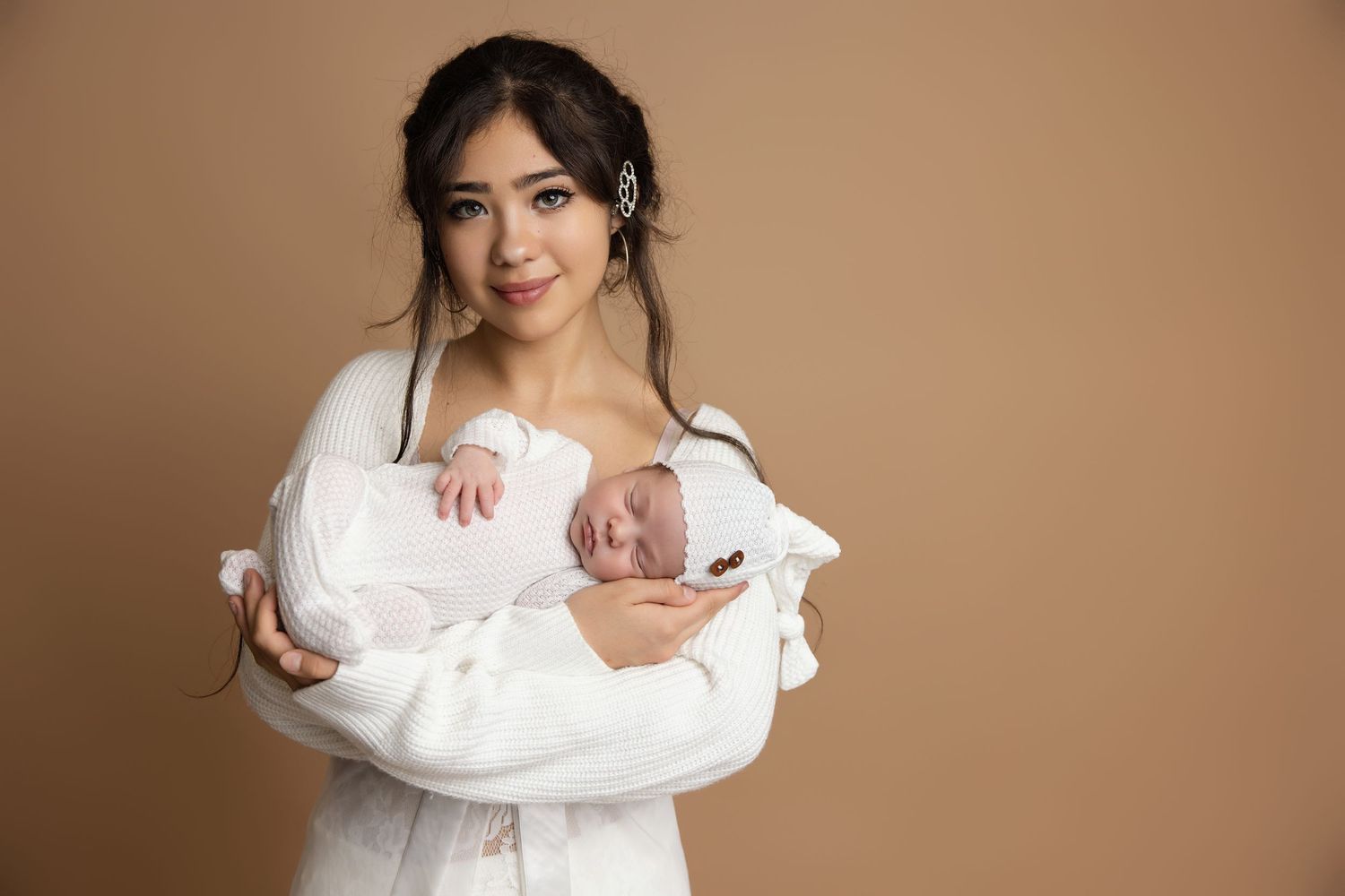 A young woman in a white outfit tenderly holds her sleeping newborn baby against a warm beige background.