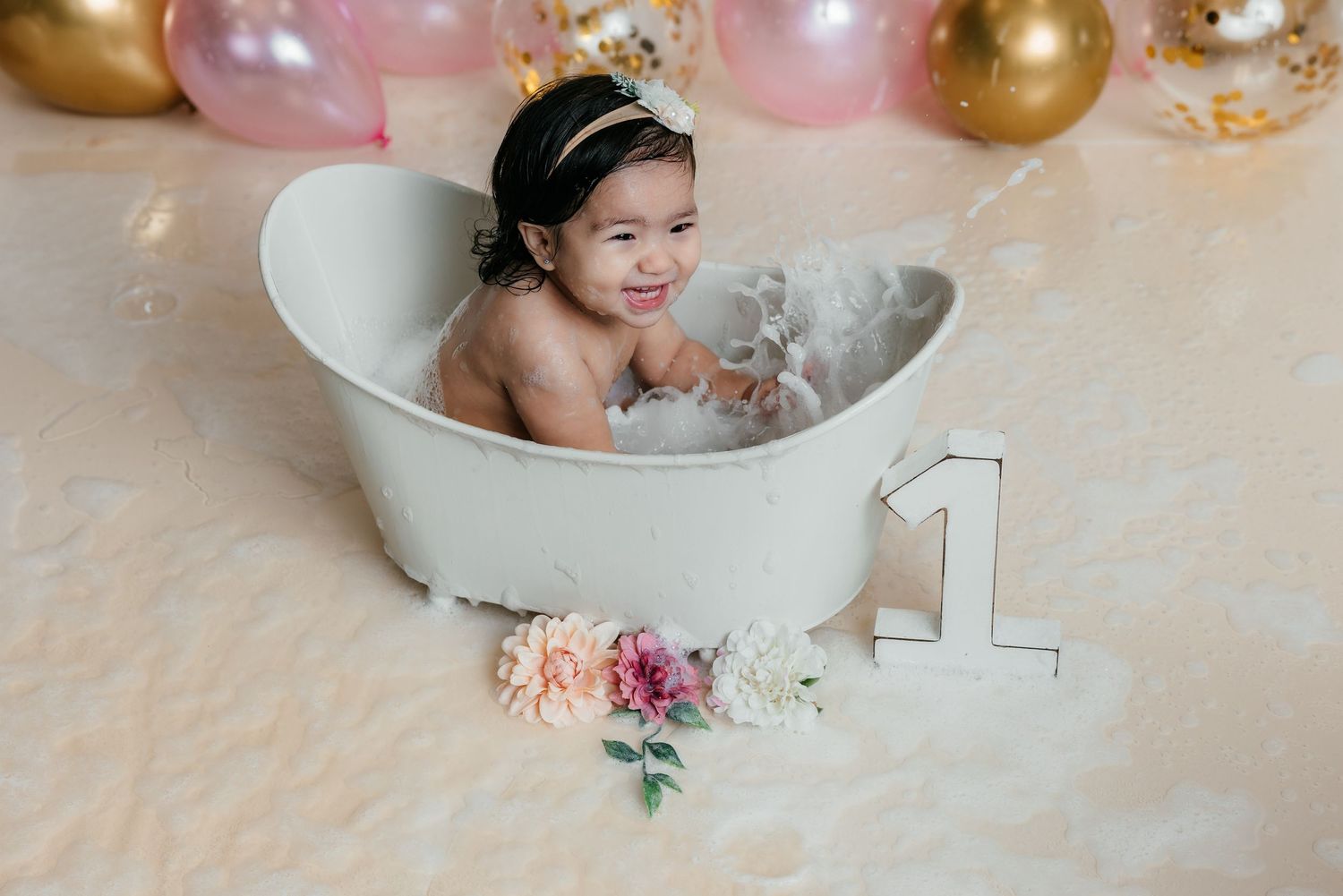 Smiling baby girl in a white bathtub with bubbles, wearing a crown headband, celebrating her first birthday.