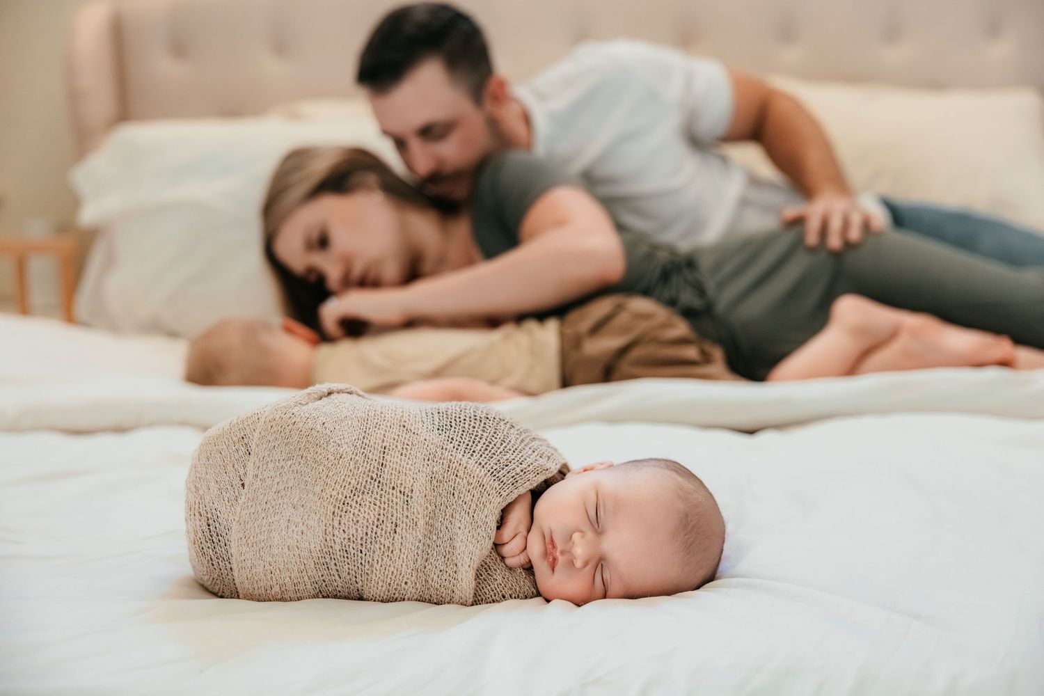 New parents lying on a white bed gazing at their peacefully sleeping swaddled newborn baby.