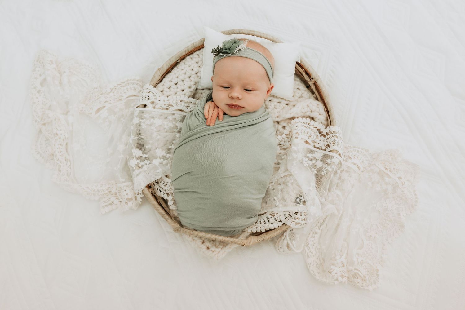 Newborn baby swaddled in sage green wrap, sleeping peacefully in a wicker basket with cream macrame blanket.