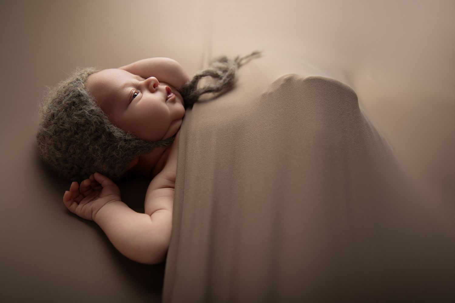 Peaceful newborn baby in a fuzzy gray knit hat sleeps against a soft taupe draped backdrop in Bonsall CA