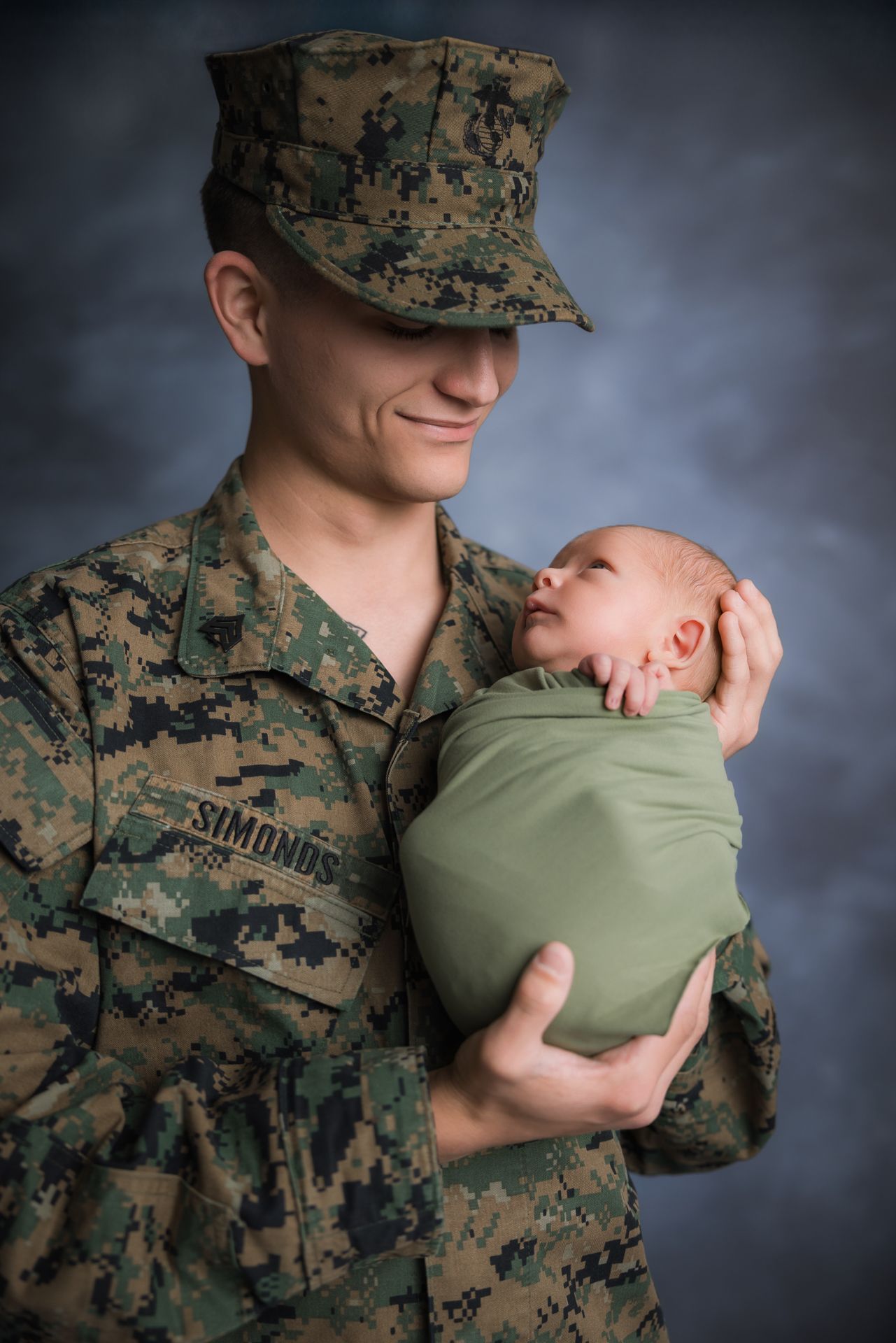 Smiling US Marine in camouflage uniform tenderly holding green swaddled newborn baby at the Studio Bonsall near Oceanside & Camp Pendleton