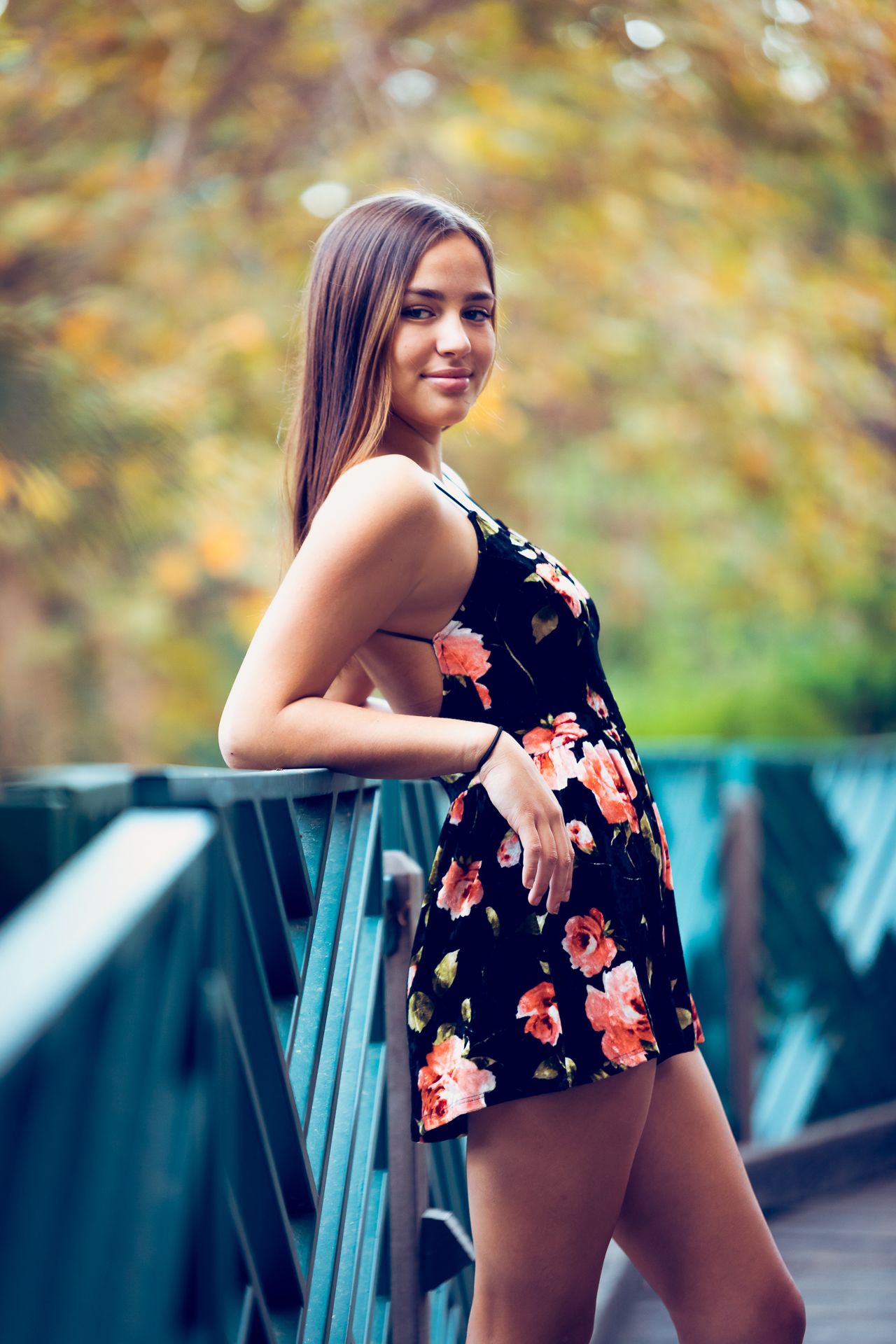 Smiling young woman in black floral dress leans on green bridge railing in autumn park.