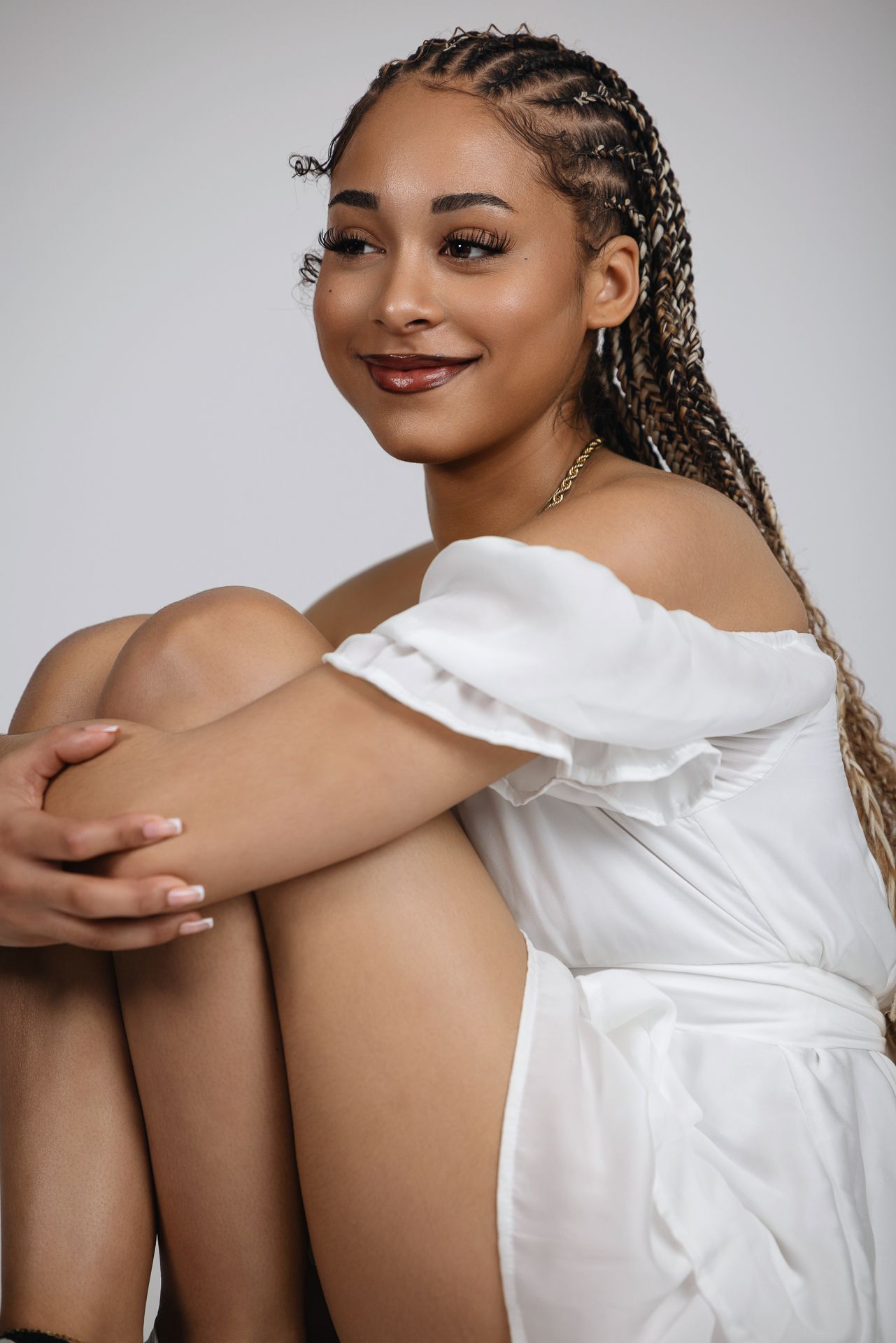 Young woman with long box braids sits hugging her knees in a white off-shoulder dress. Senior photography session in Bonasll near Vista
