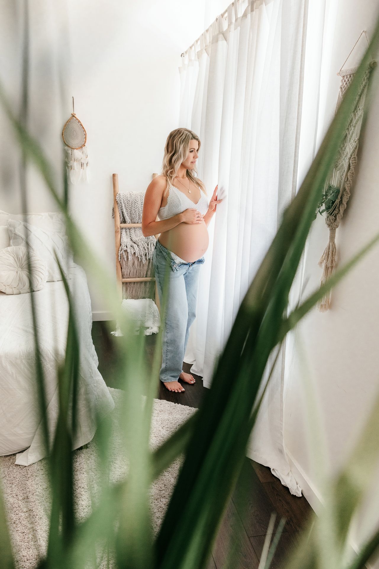 Pregnant woman in jeans and white top standing by curtained window with green plants in foreground in the Bonsall Studio near Temecula