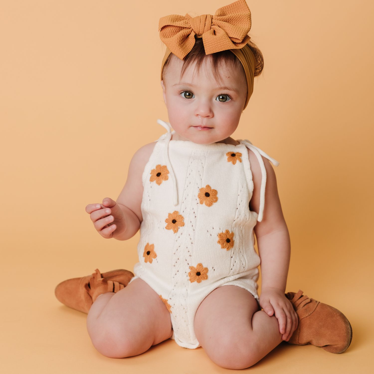 Adorable baby girl in floral knit romper and bow headband on yellow background.