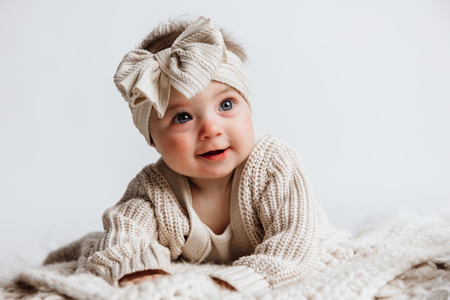 Adorable baby in beige knit sweater and bow headband smiling while lying on a soft blanket.