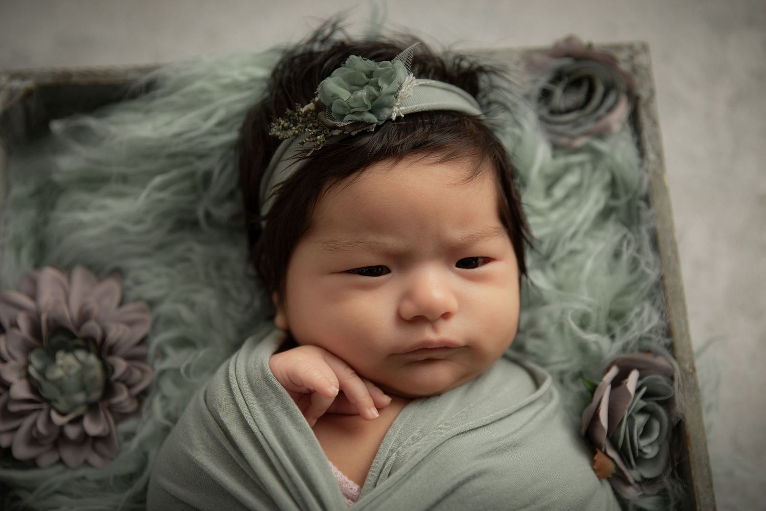 Newborn baby wrapped in sage green swaddle with floral headband, resting in a decorated wooden box.