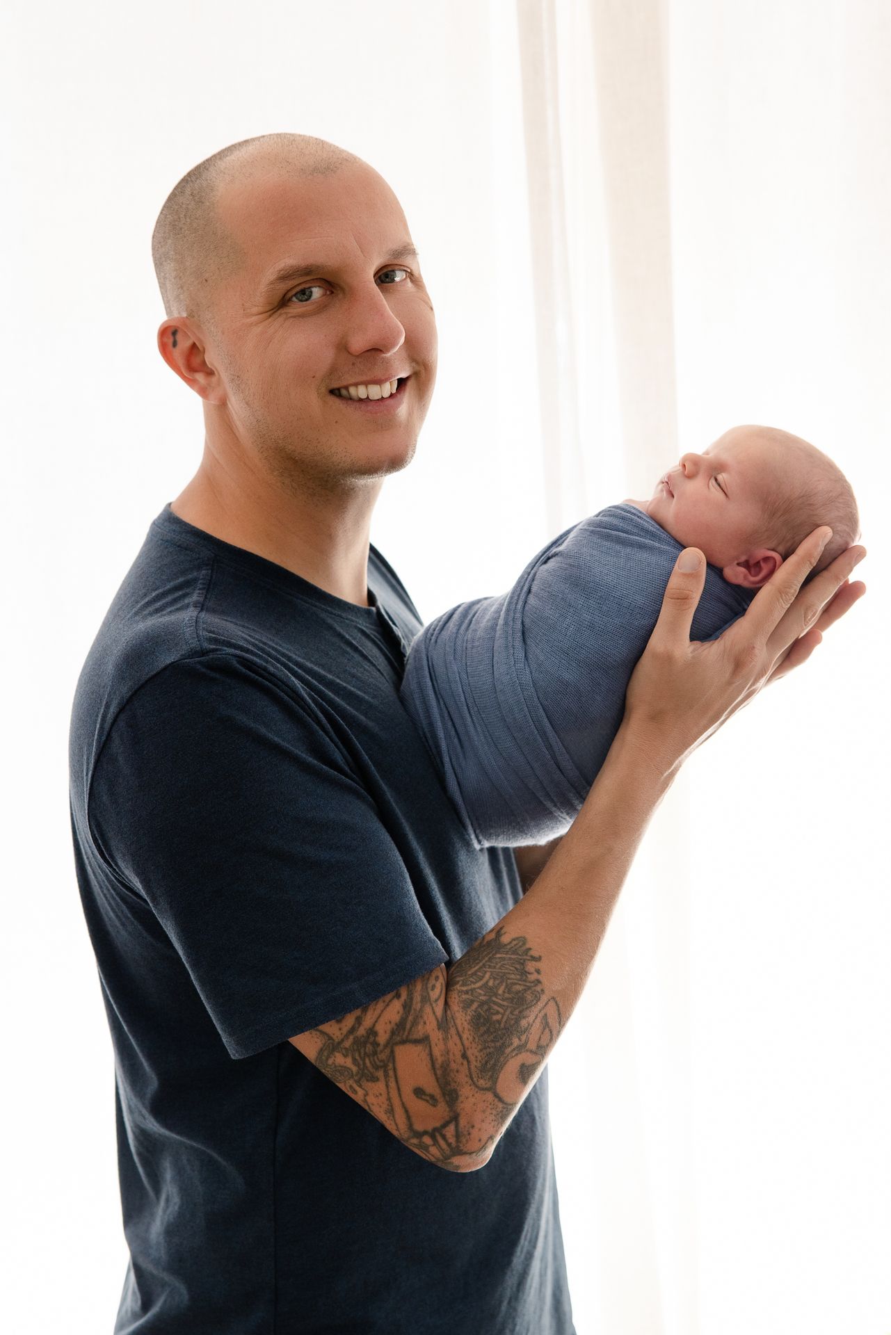 Smiling bald man with tattoos holding a sleeping newborn baby wrapped in a blue swaddle blanket.
