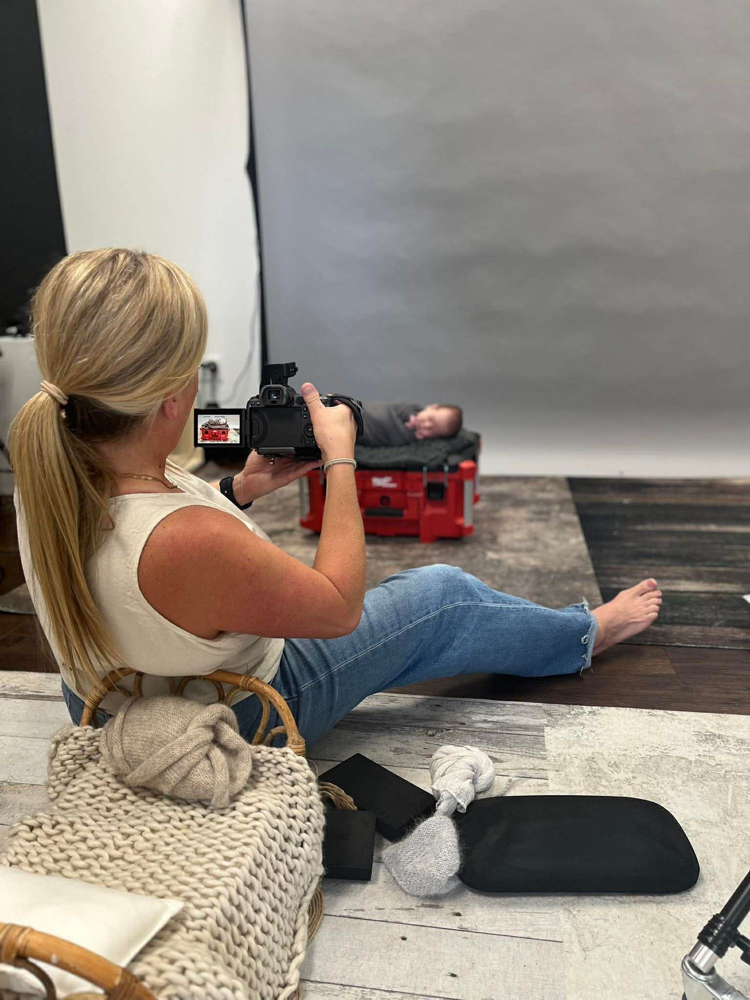 Blonde female photographer sitting on floor in studio, photographing a newborn baby on a red posing bed.