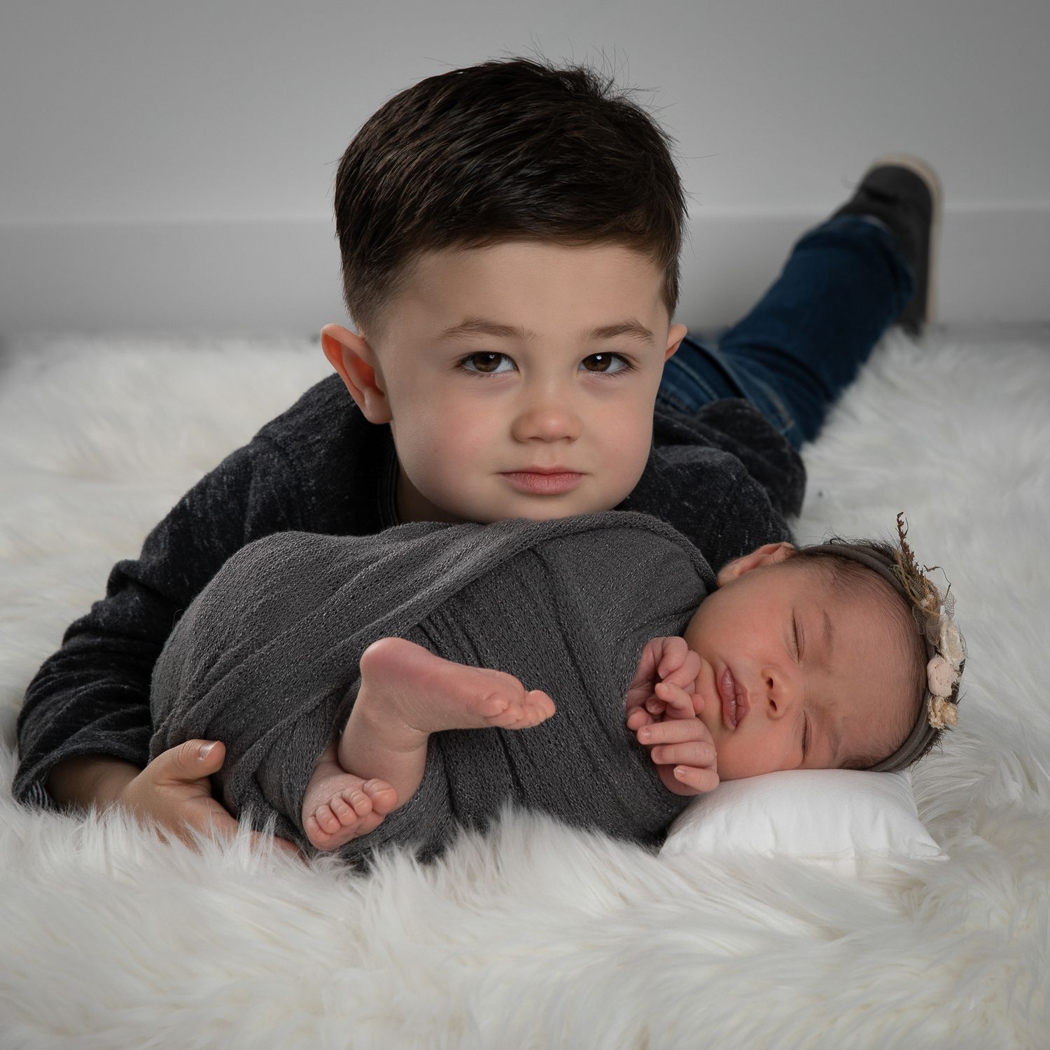 A young toddler boy lies on a white fur rug cradling a sleeping newborn baby wrapped in a gray swaddle.