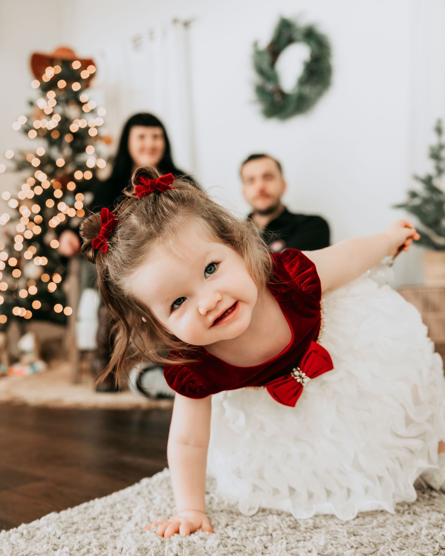 A smiling toddler girl in a red and white dress crawls toward the camera with a Christmas tree and parents in the background.