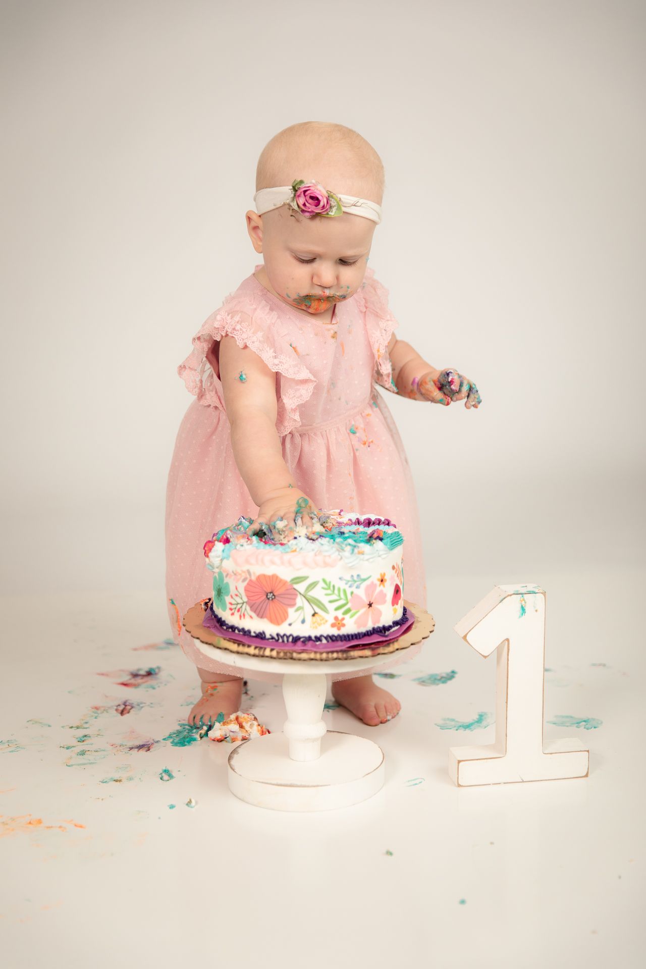 Baby girl in pink dress touching a decorated first birthday cake with number one sign beside it.