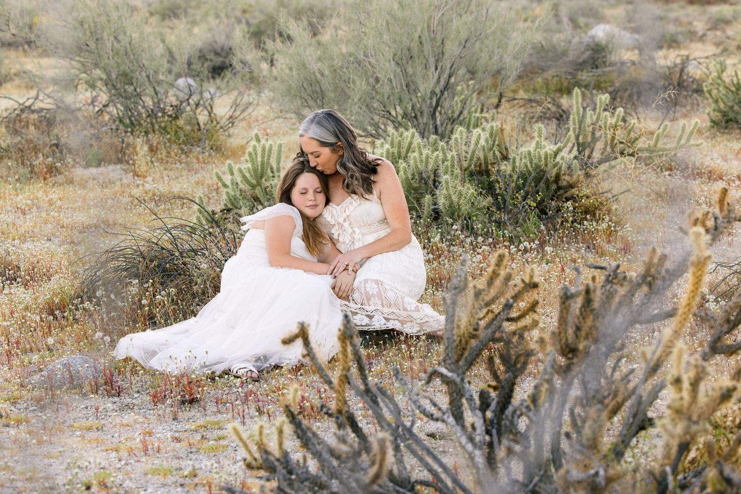 Two brides in white dresses sitting together among desert shrubs and cacti in a serene outdoor setting.