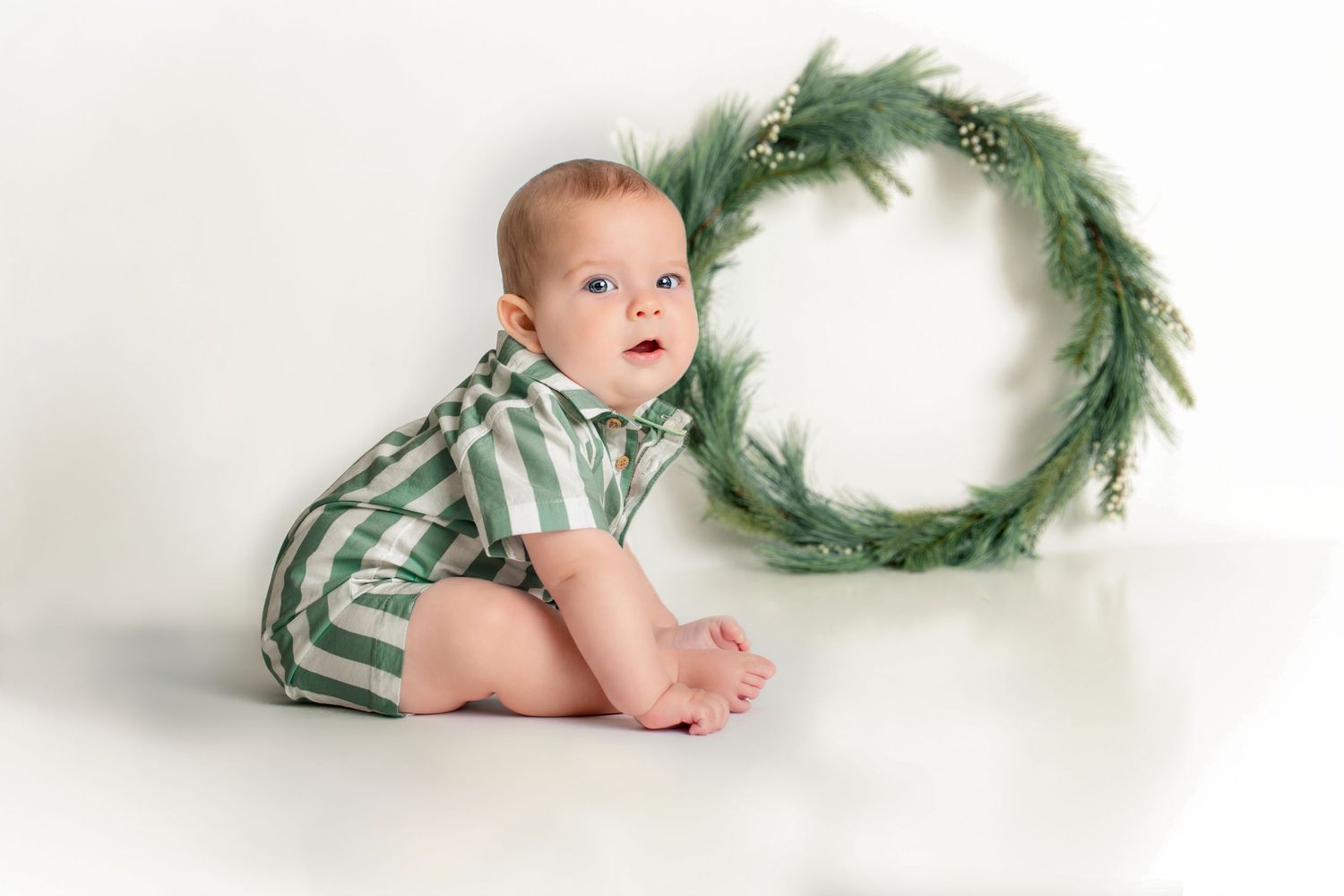 Baby in green striped romper sits beside a festive wreath on white background.