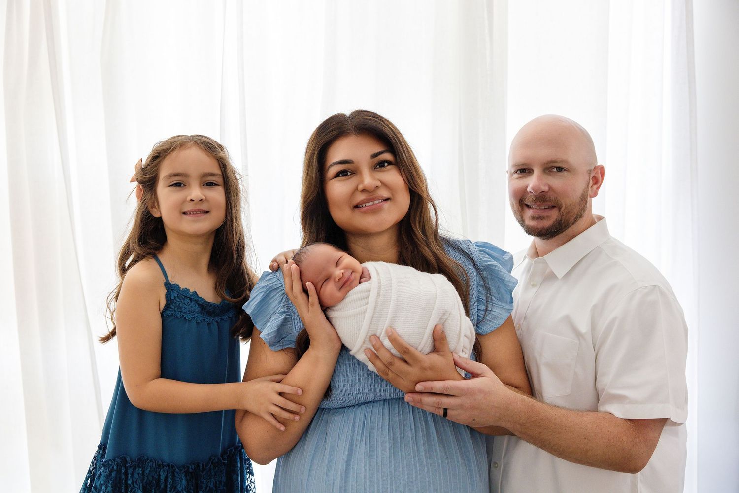 Family of four posing indoors, mother holding newborn baby, father and young daughter smiling, white curtain background.