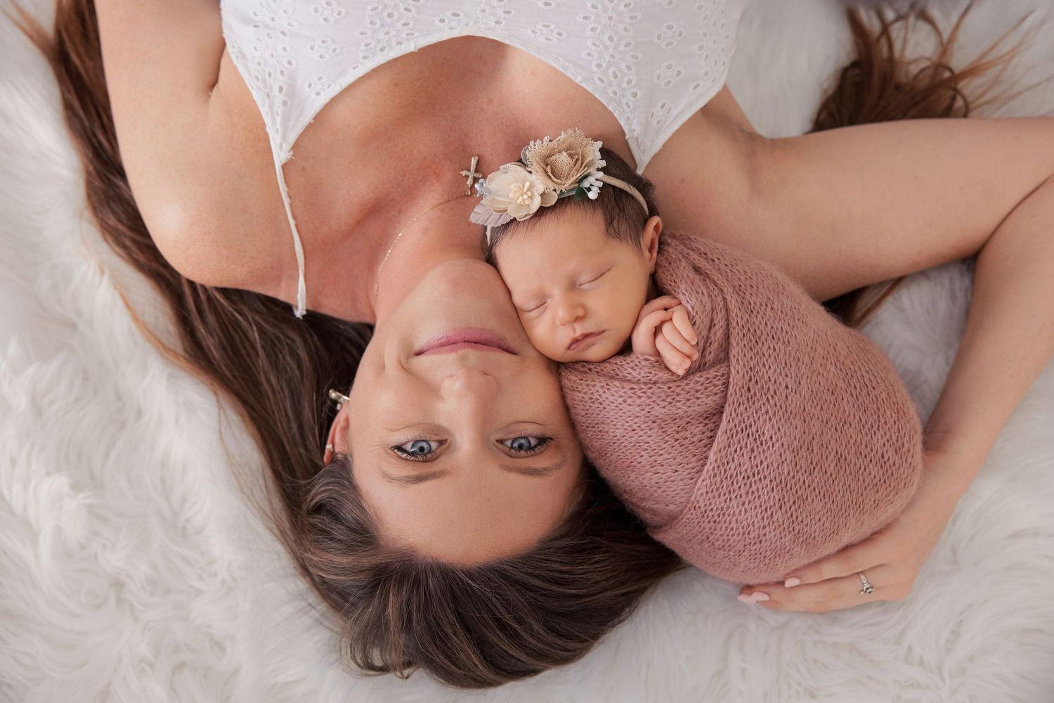 Mother lying down with newborn baby wrapped in pink swaddle and floral headband on white fur backdrop.