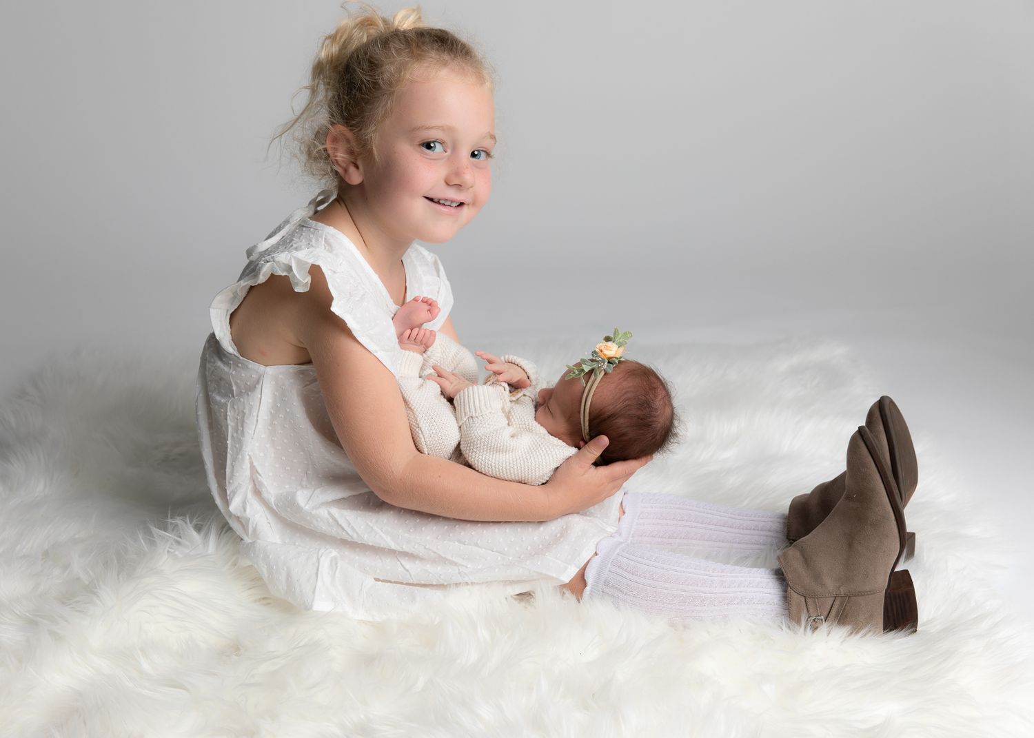 Young girl in white dress smiling while holding a newborn baby on a fluffy white rug in a studio.