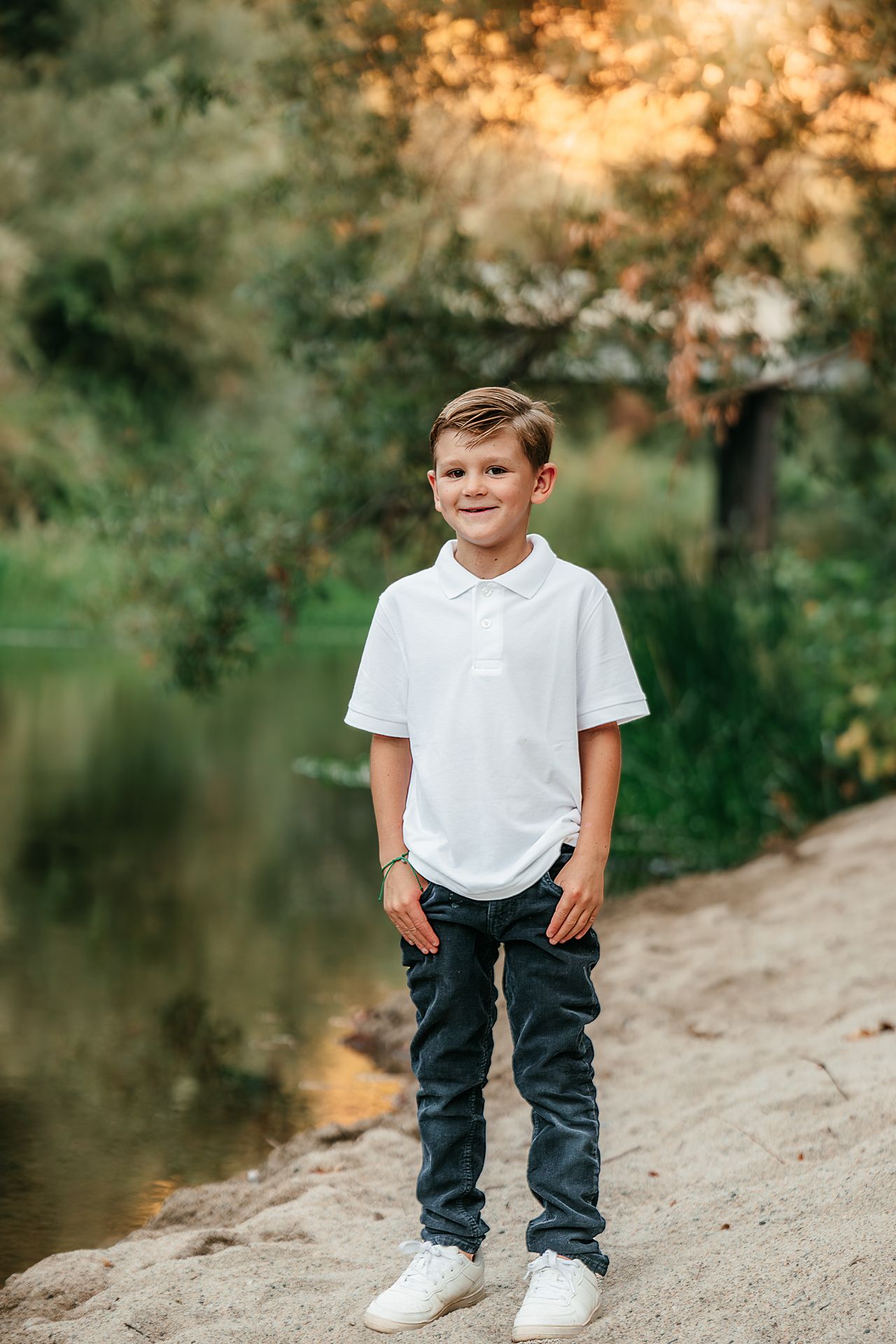 Smiling young boy in a white polo shirt standing near a peaceful riverside at golden hour.