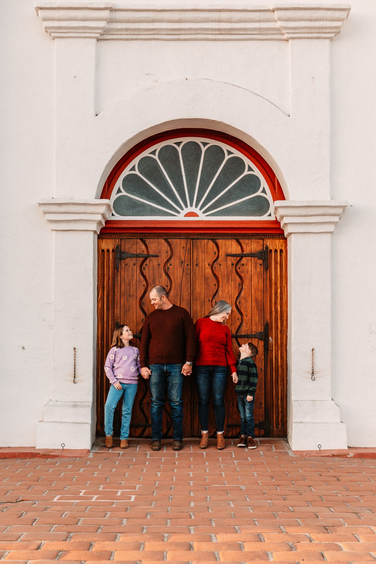 Family of four poses in front of ornate wooden church doors with arched entryway in Oceanside near bonsall