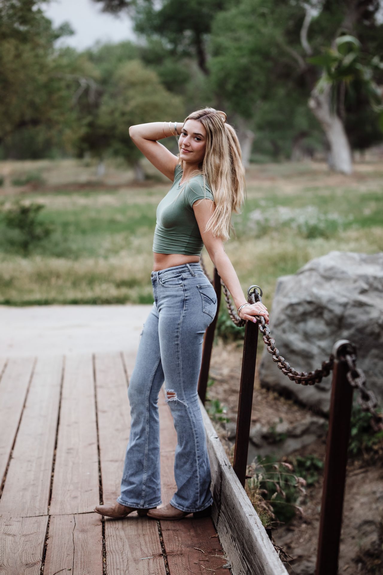 Blonde woman in green crop top and flared jeans posing on wooden deck in park.