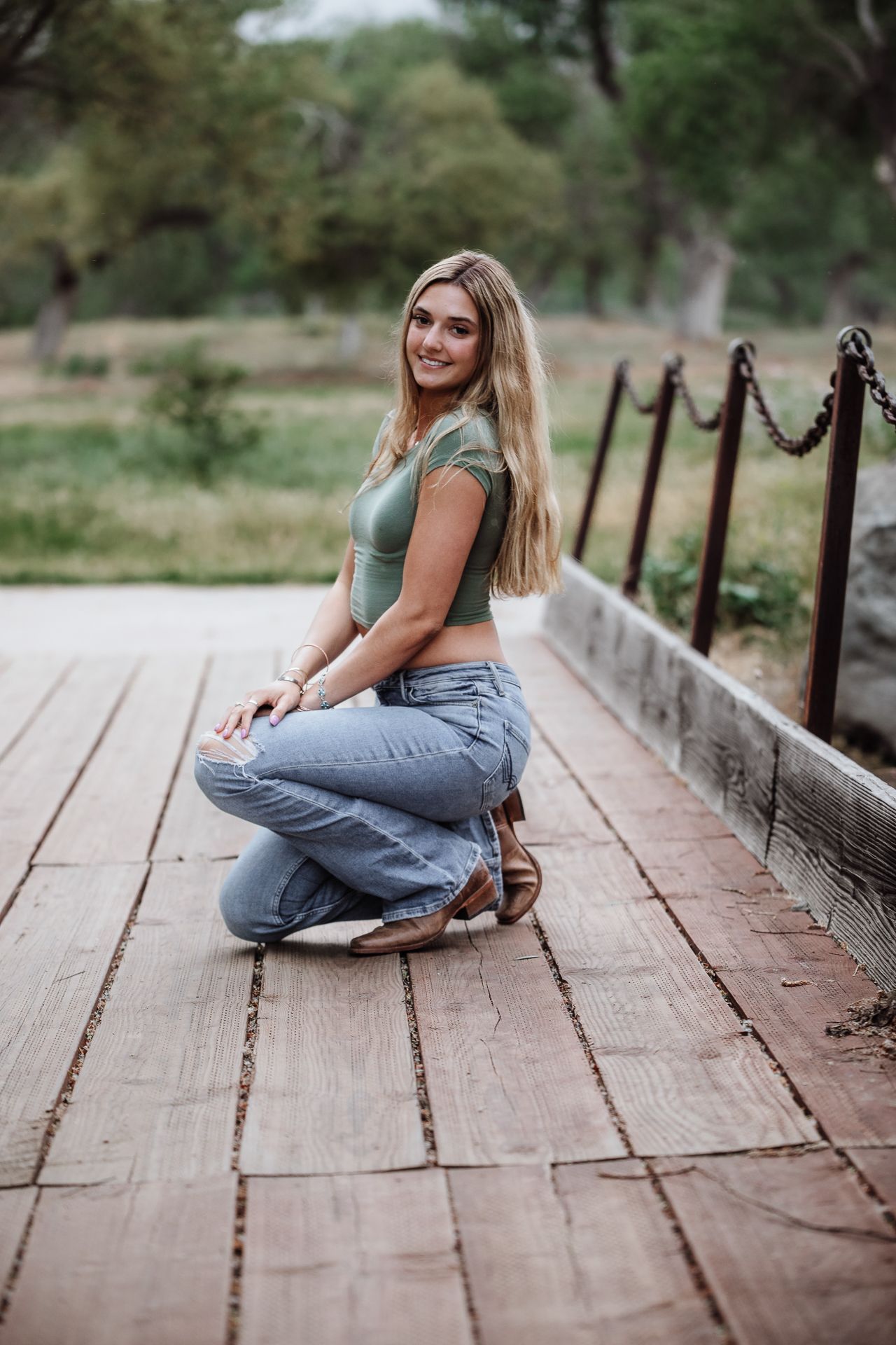 Smiling blonde girl in green crop top and jeans crouching on wooden bridge for senior portrait outdoors.