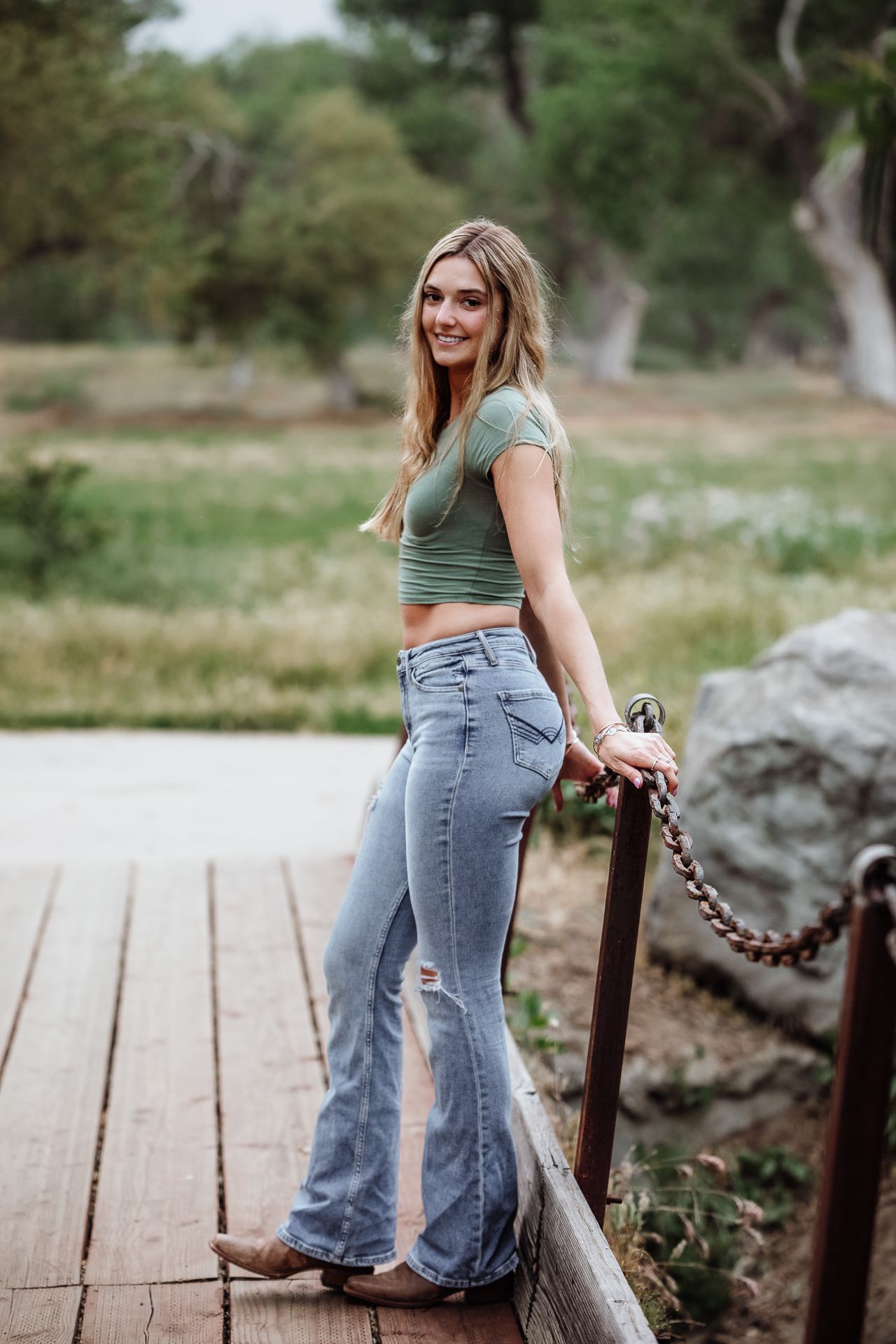 Young blonde woman in green crop top and flare jeans posing on wooden bridge in outdoor nature setting.