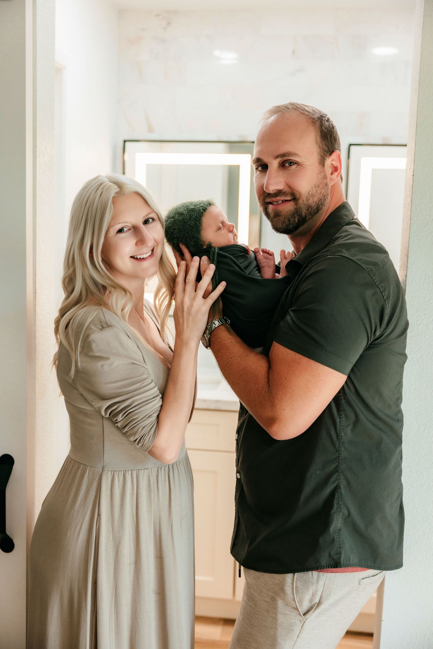 A smiling couple poses indoors holding their newborn baby, the woman in a sage dress and man in a dark green shirt.