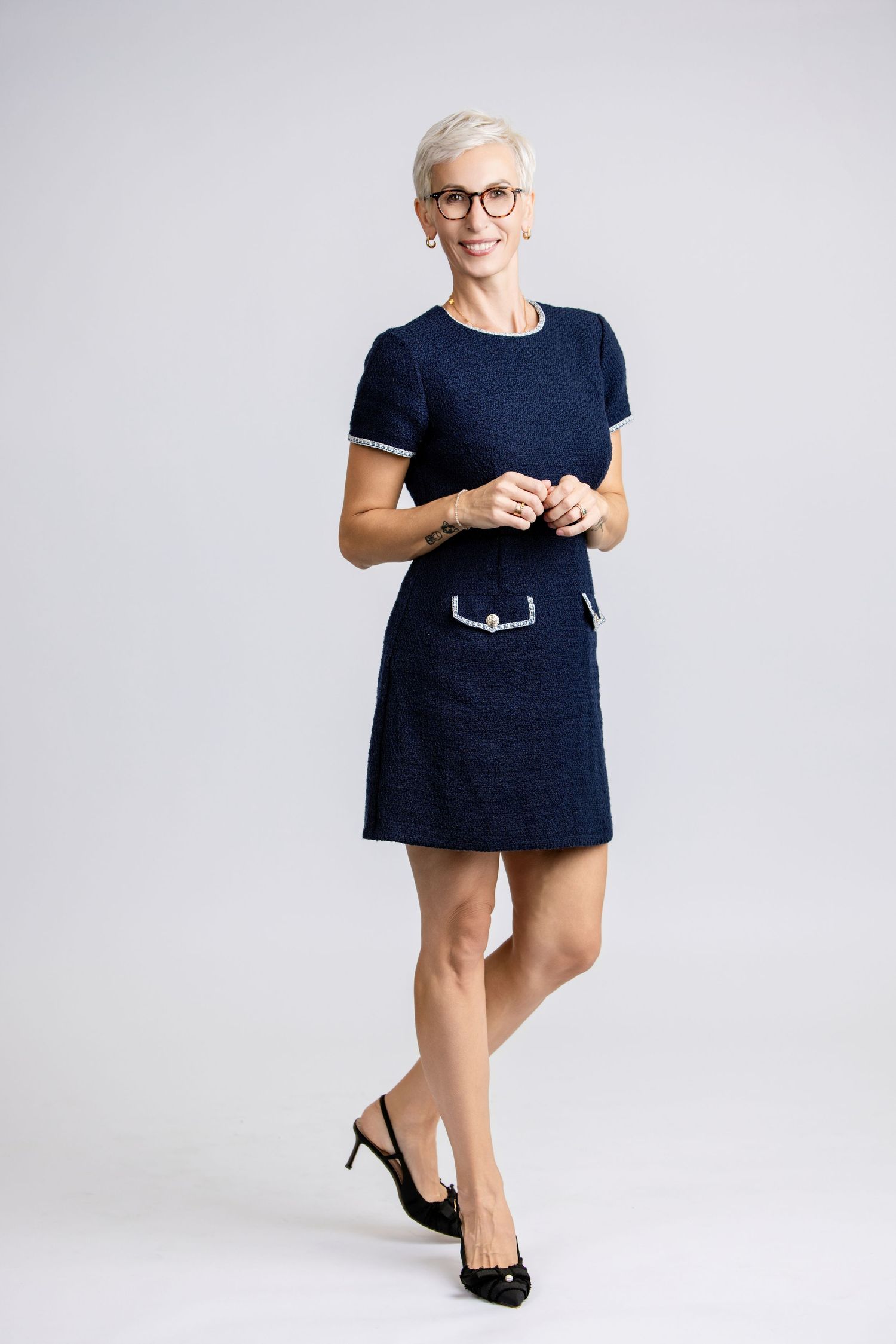 Woman in navy blue tweed mini dress with glasses and black heels posing against gray background.
