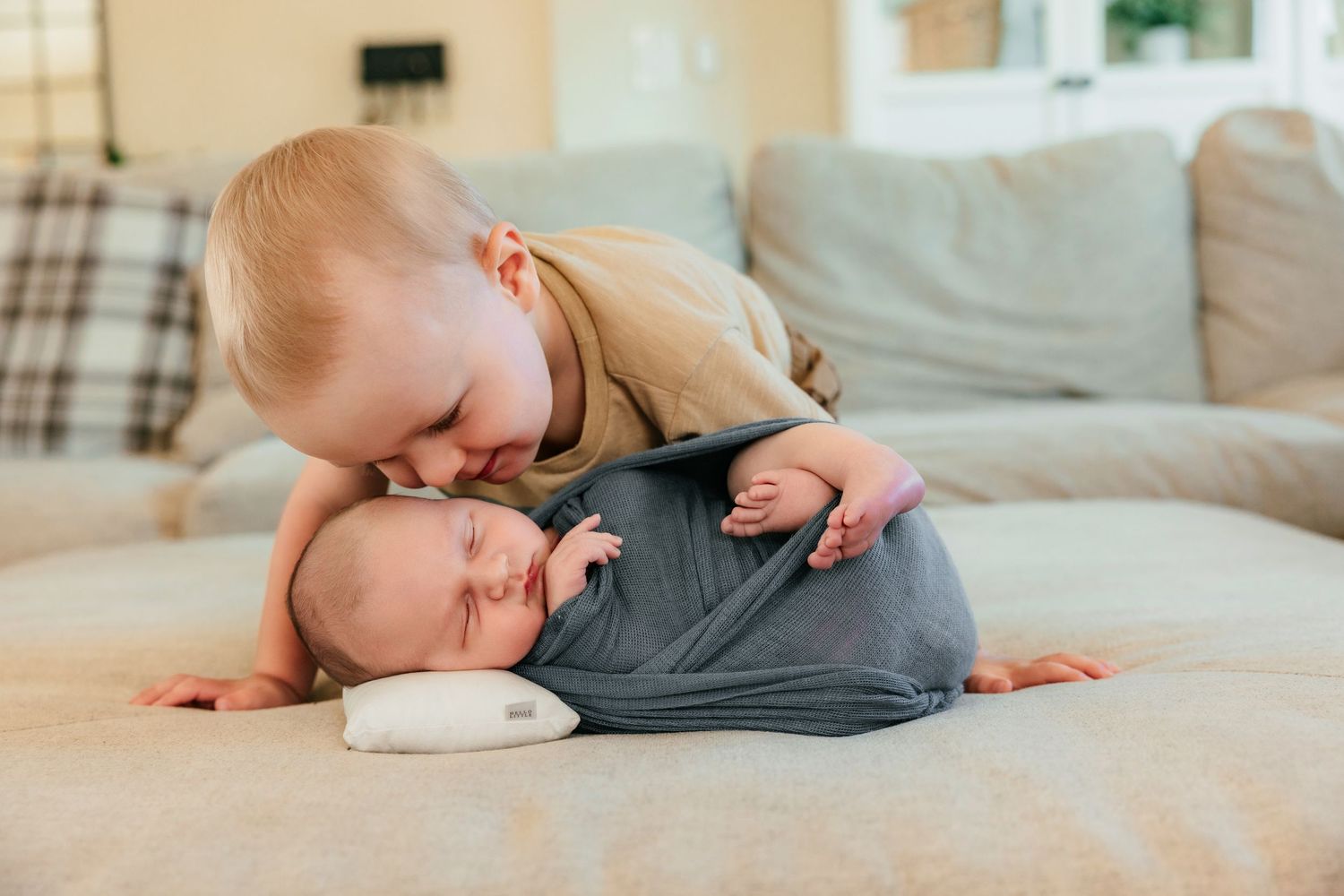Toddler gently kissing sleeping newborn baby sibling wrapped in gray swaddle on couch.