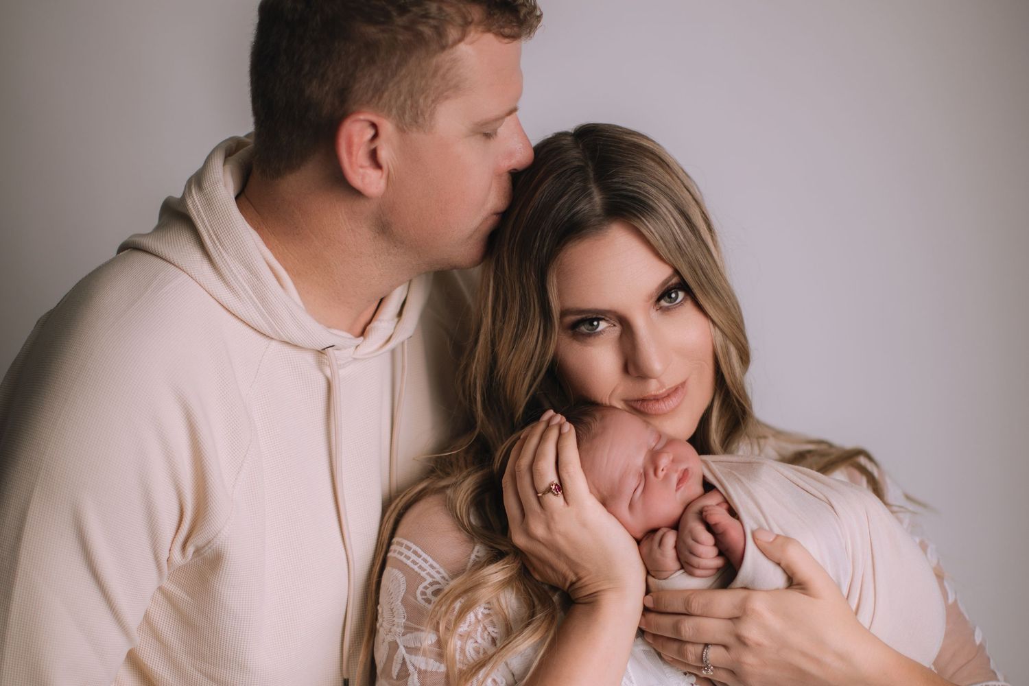 A couple tenderly holds their newborn baby, the man kissing the woman's head, in a soft studio portrait.