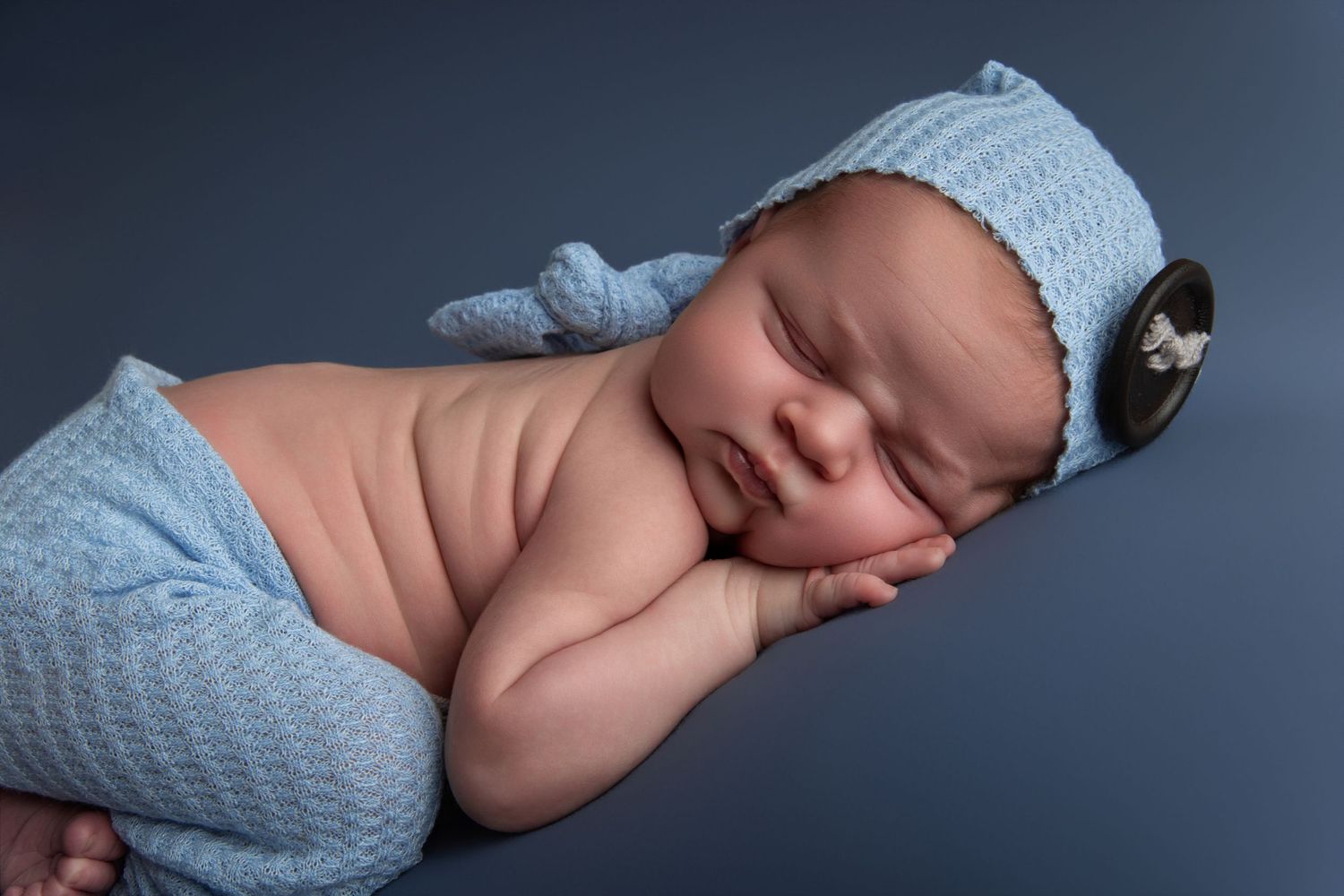 Newborn baby sleeping peacefully in blue knit hat and wrap on gray background.