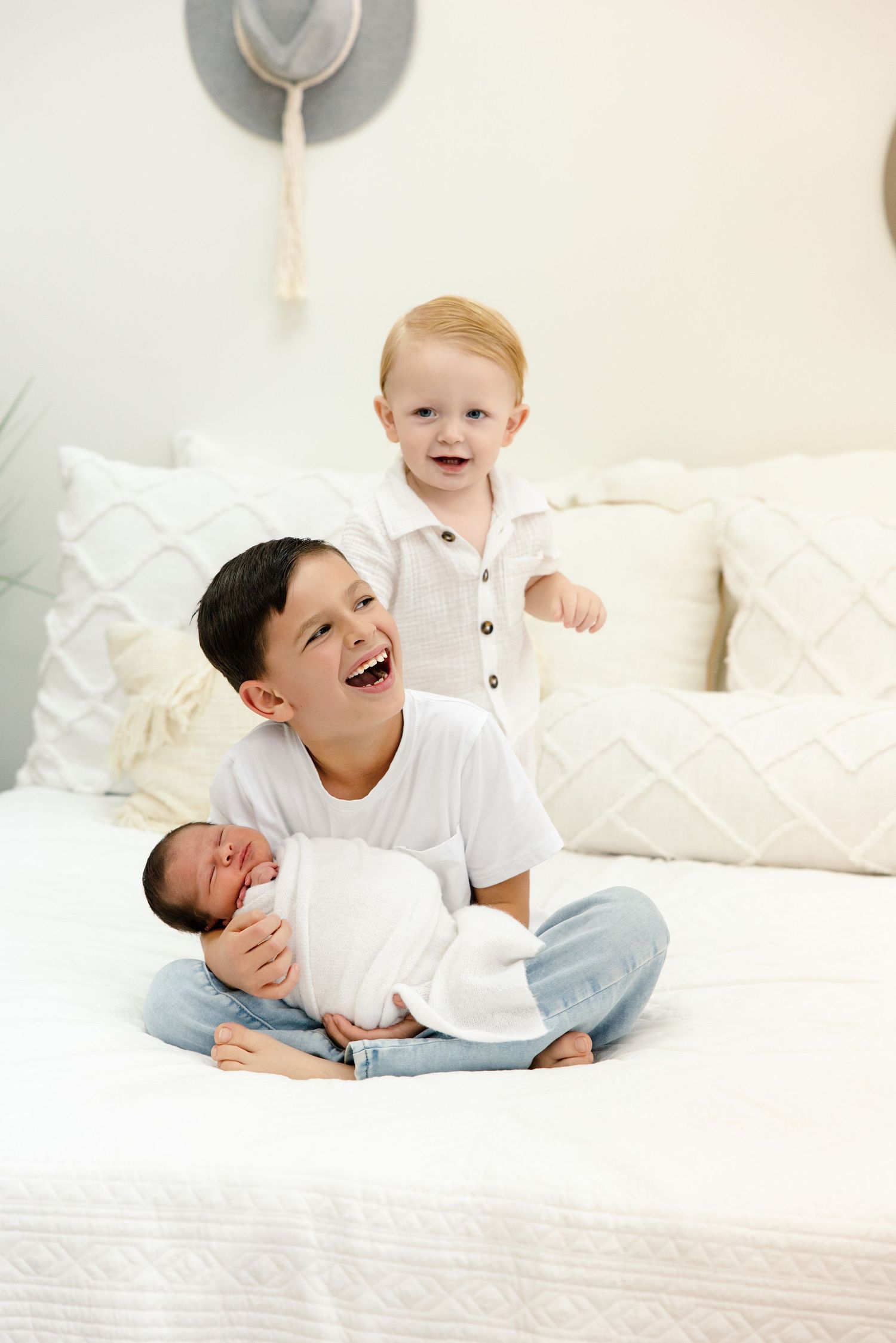 Three brothers sit on a white bed, oldest holding newborn baby, middle toddler standing behind, all smiling joyfully.