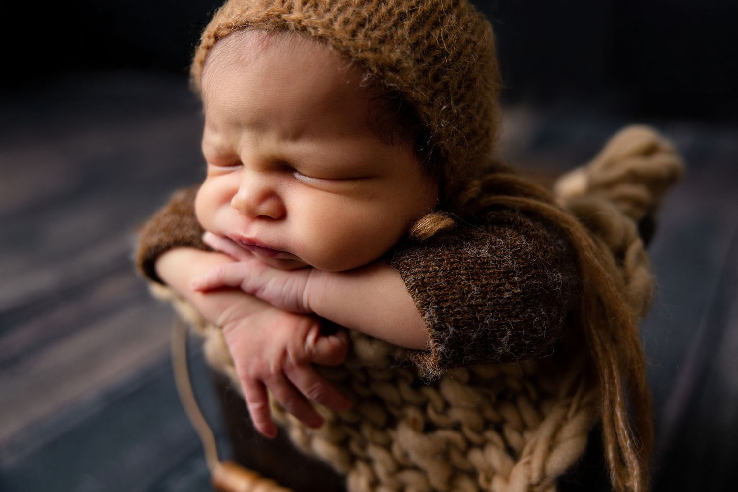 Sleeping newborn baby in brown knit bonnet and outfit posed with hands under chin in cozy studio portrait.