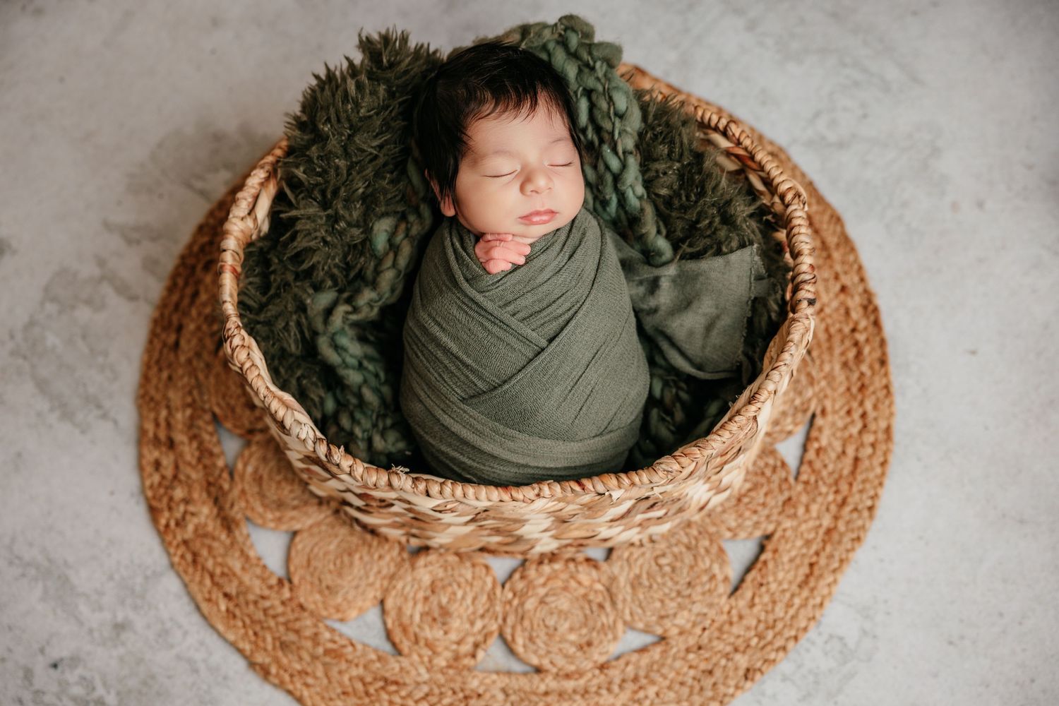 Newborn baby swaddled in green wrap sleeping peacefully in a woven basket with green knit blanket.