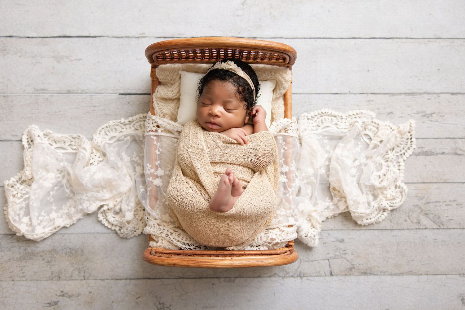 Newborn baby swaddled in cream wrap with lace bonnet sleeping in a tiny wooden bed on white wood floor.