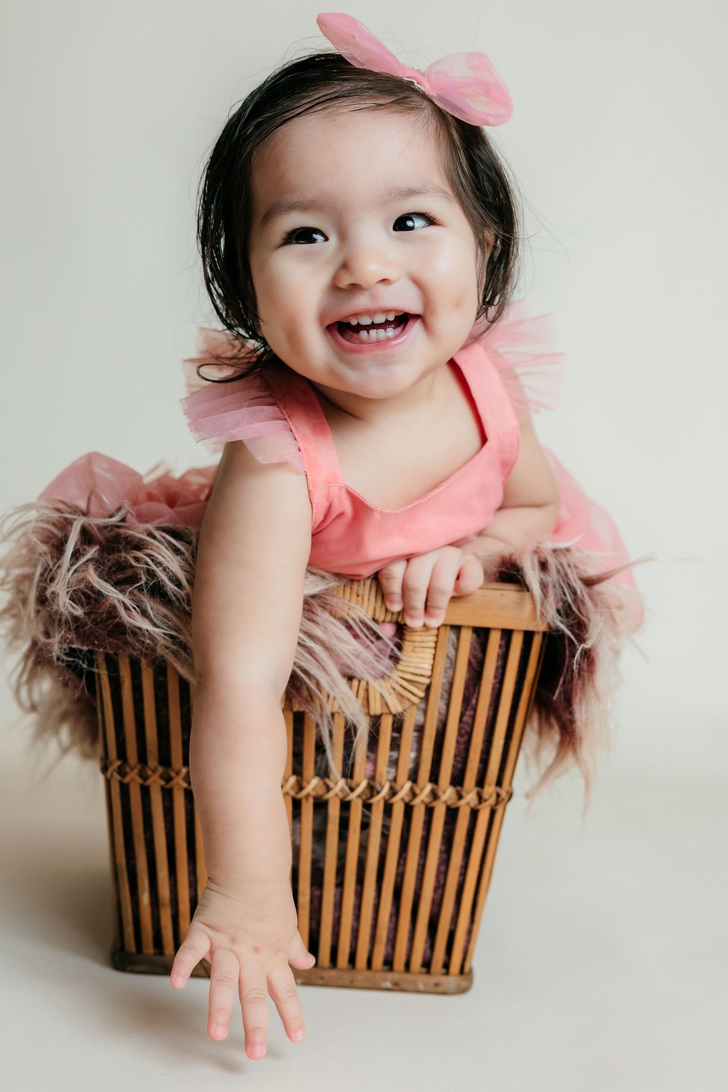 Smiling baby girl in pink tutu sitting inside a wicker basket during a studio photo session.