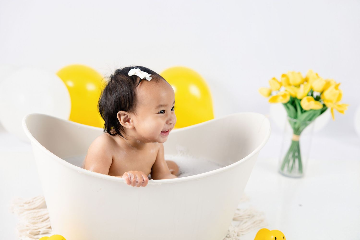 Happy baby sitting in a white bathtub surrounded by yellow balloons and rubber ducks in a bright white studio.