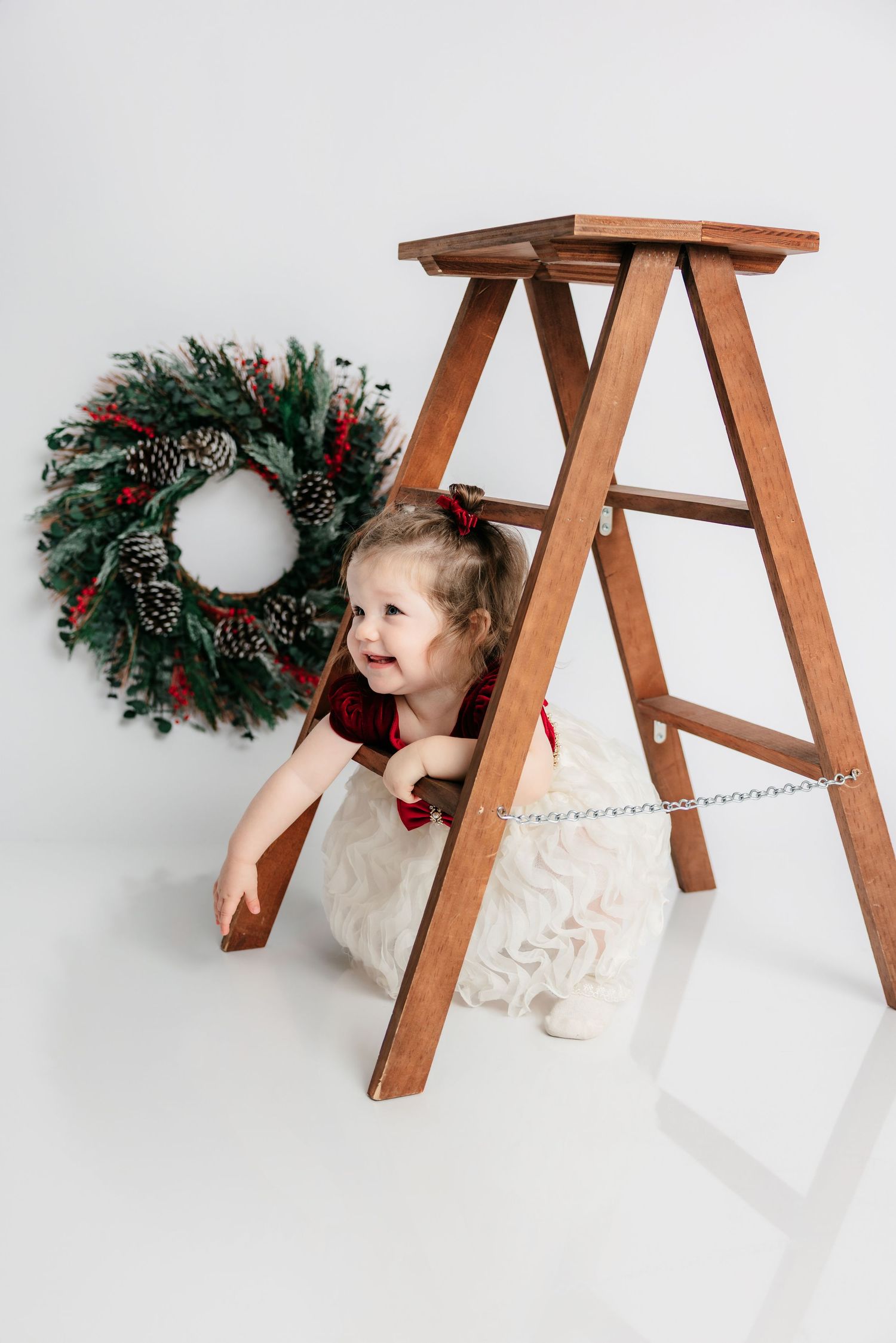 A cheerful toddler girl in a white dress crouches under a wooden ladder beside a festive Christmas wreath on a white background.