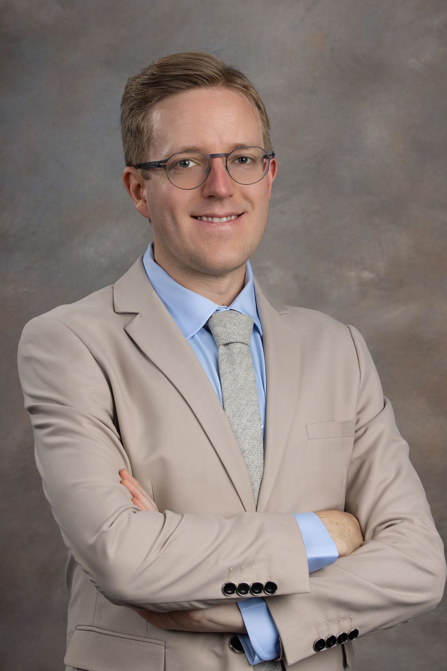 Professional man in beige suit and glasses smiling with arms crossed against neutral gray background.