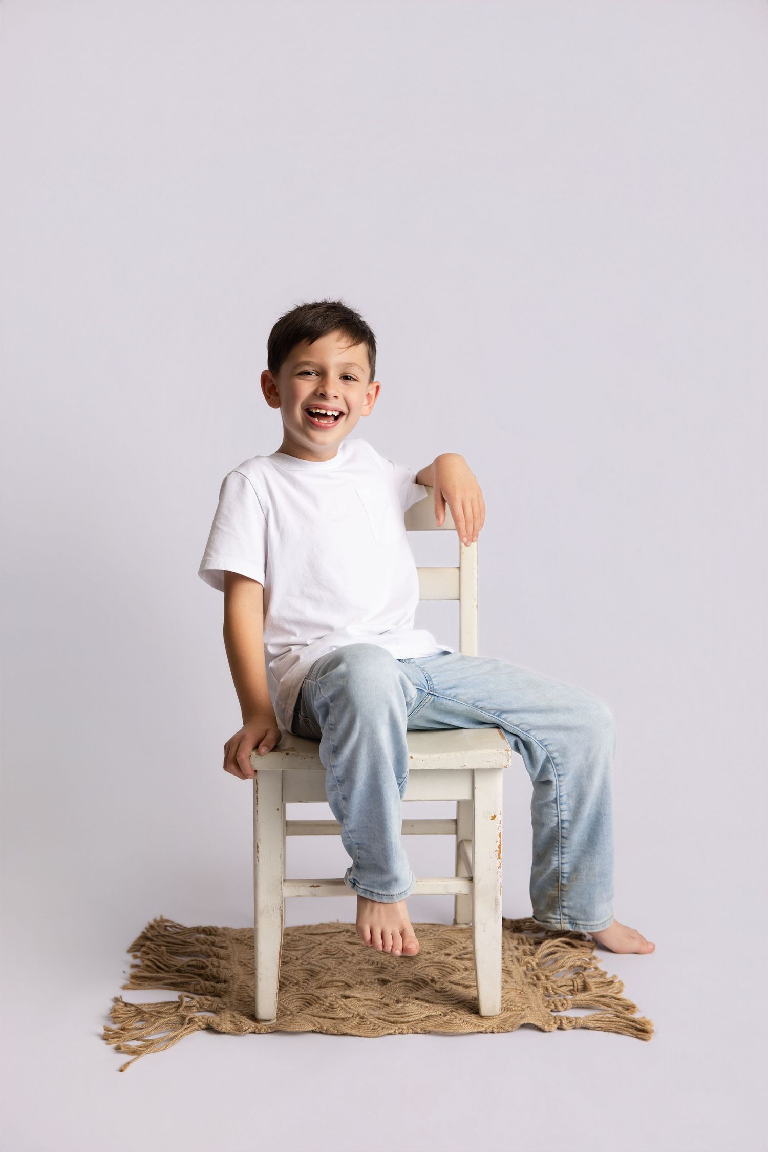A cheerful young boy in a white t-shirt and jeans sits barefoot on a white wooden chair, laughing against a gray background.
