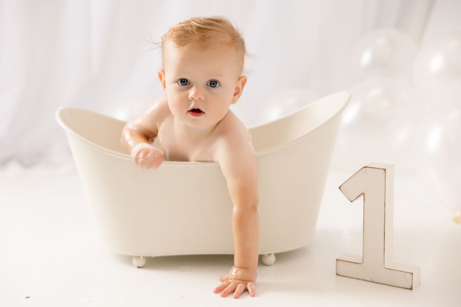 Baby sitting in a white mini bathtub next to a number one sign, celebrating their first birthday.