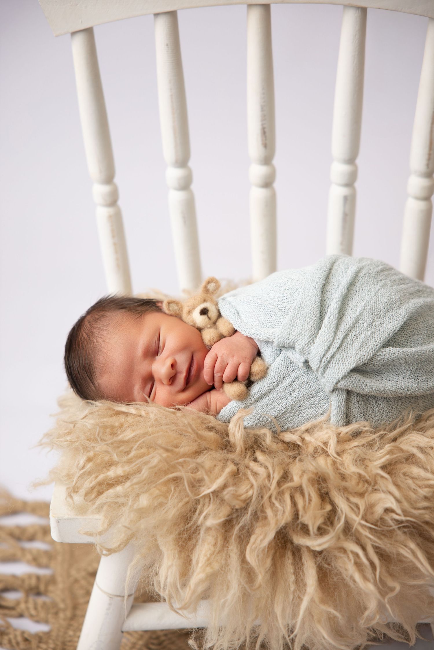 Sleeping newborn baby swaddled in blue wrap on fluffy fur blanket in white chair with teddy bear.