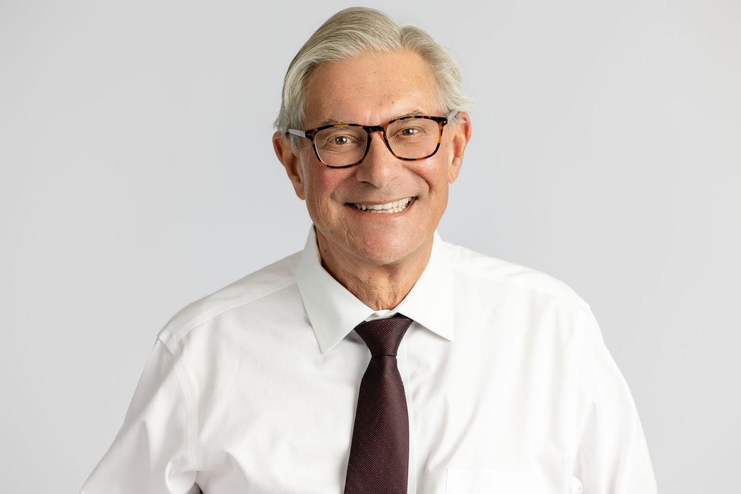 Smiling senior businessman with gray hair and glasses wearing a white shirt and dark tie against a gray background.