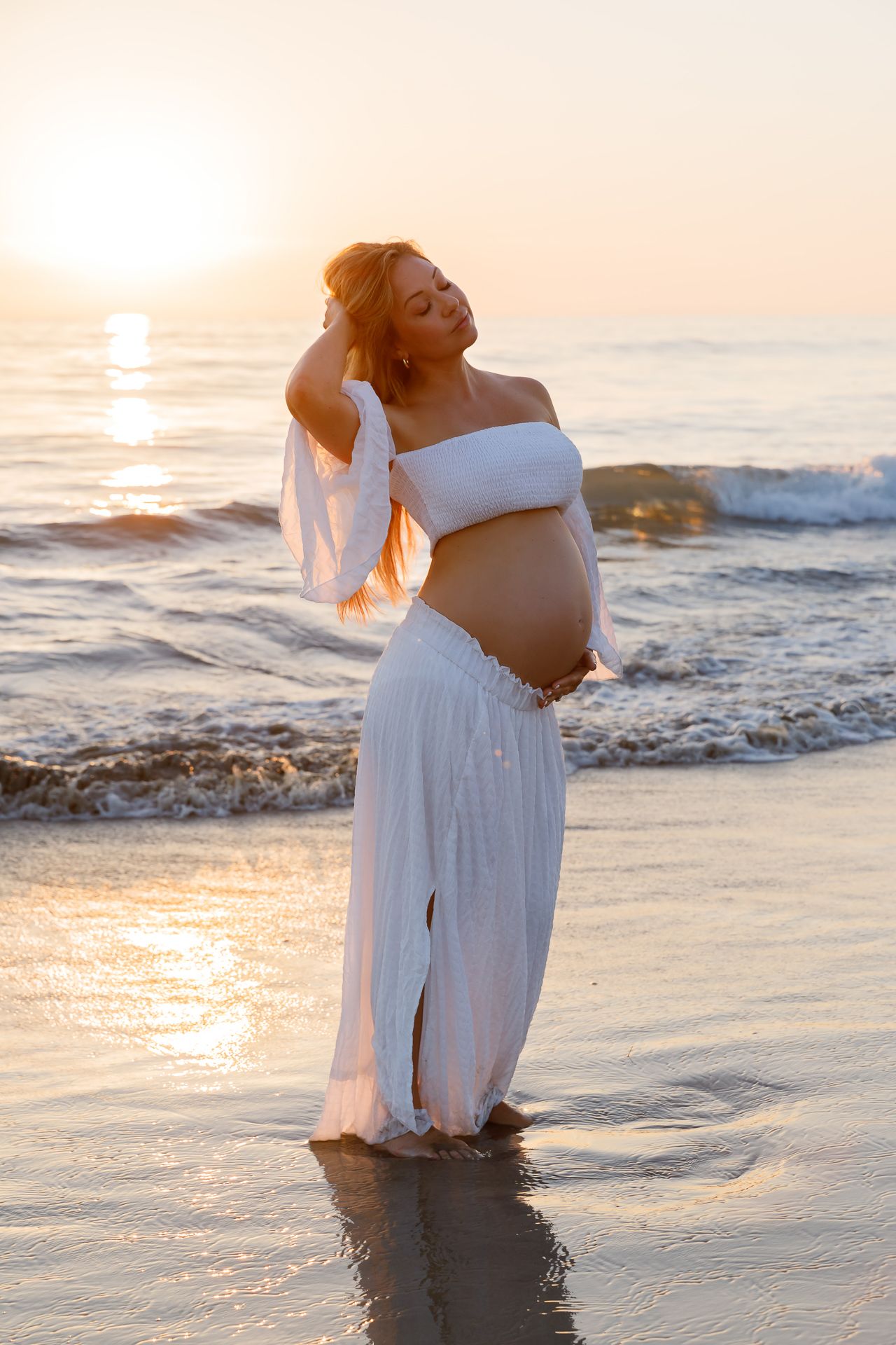 Maternity photoshoot at sunset on a beach with waves crashing and golden light reflecting off the water.