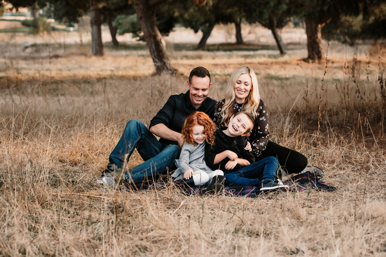 A happy family of four sits together laughing in a dry grass field during fall.