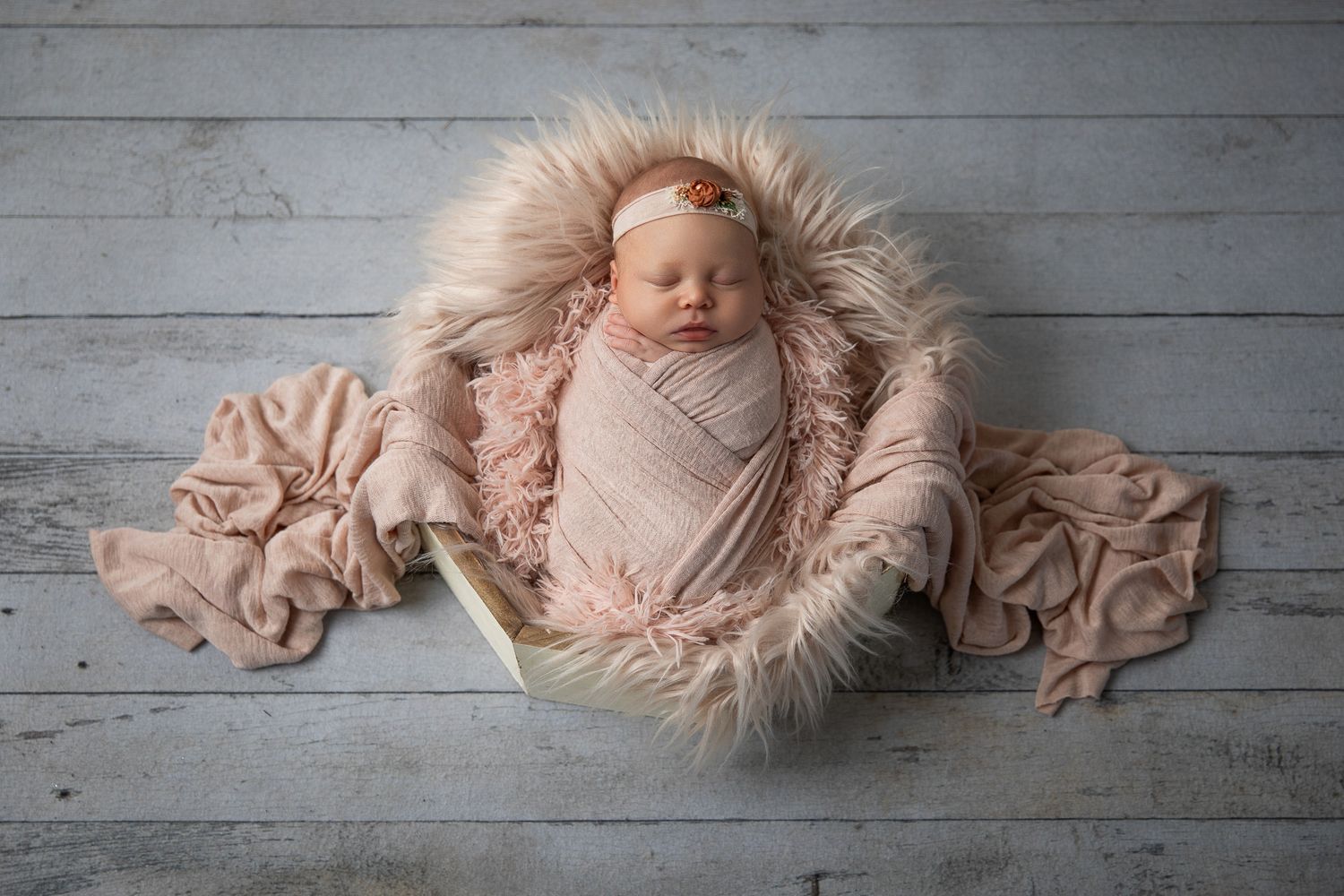 Swaddled newborn baby girl in blush pink wrap resting in a bowl with fluffy fur on wood floor.