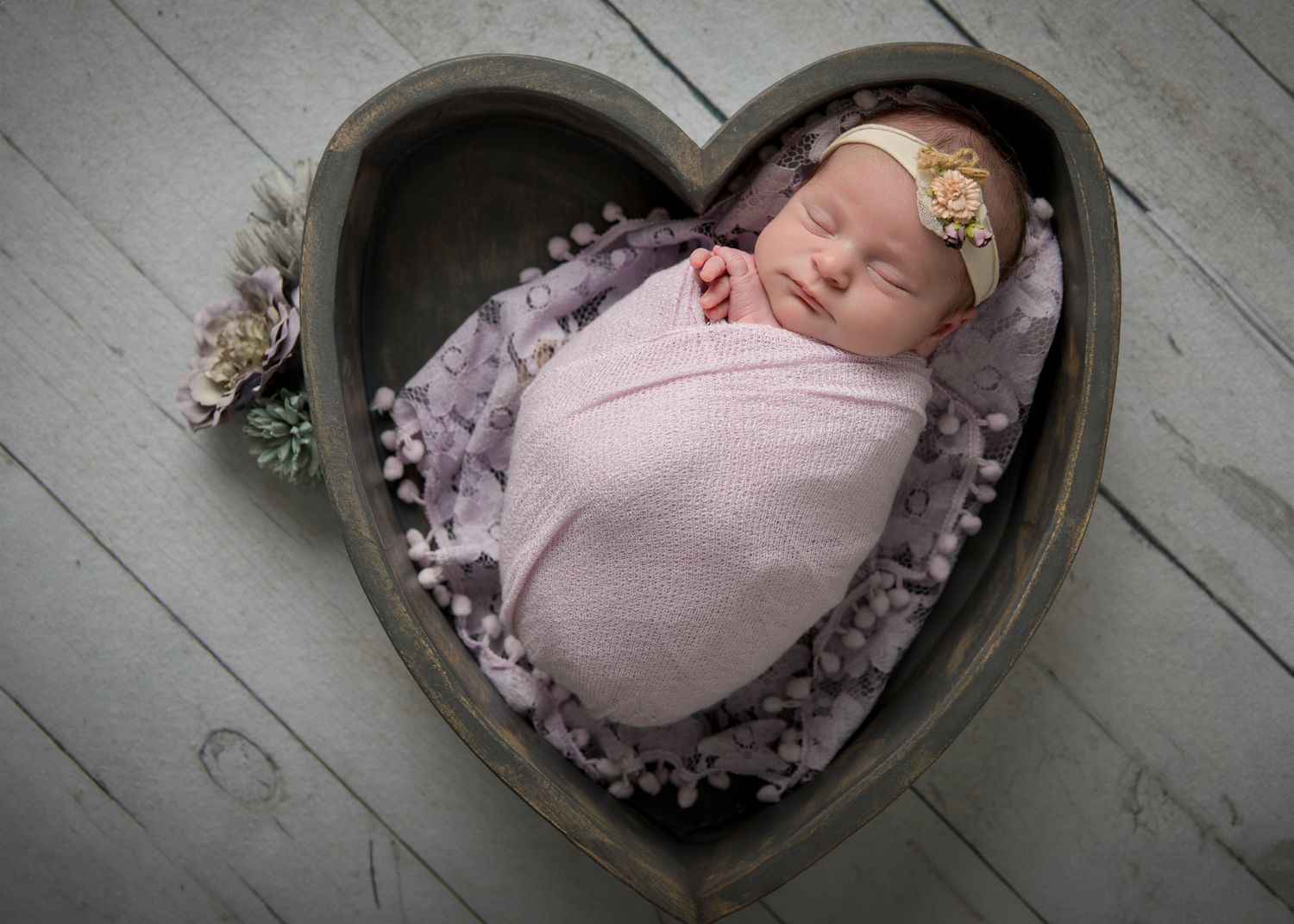 Newborn baby girl swaddled in pink wrap sleeping in heart-shaped wooden bowl with floral headband.