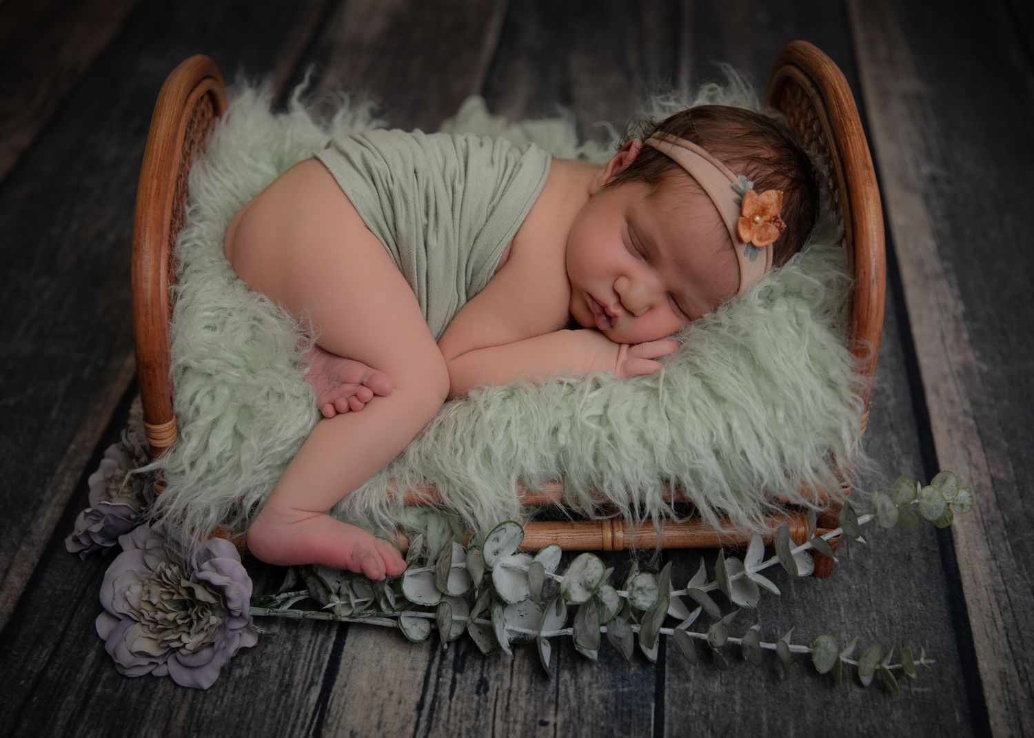 Newborn baby sleeping peacefully on a fluffy mint green fur blanket in a small wooden bed with floral decorations.