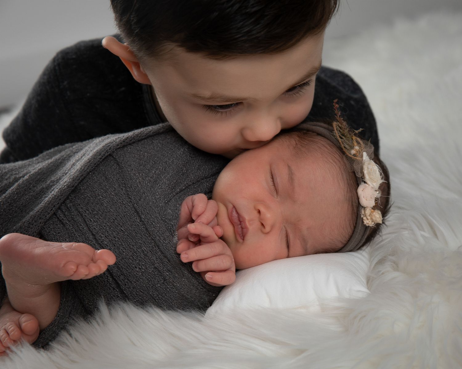 Older boy gently kissing sleeping newborn baby wrapped in gray swaddle with floral headband on white fur.